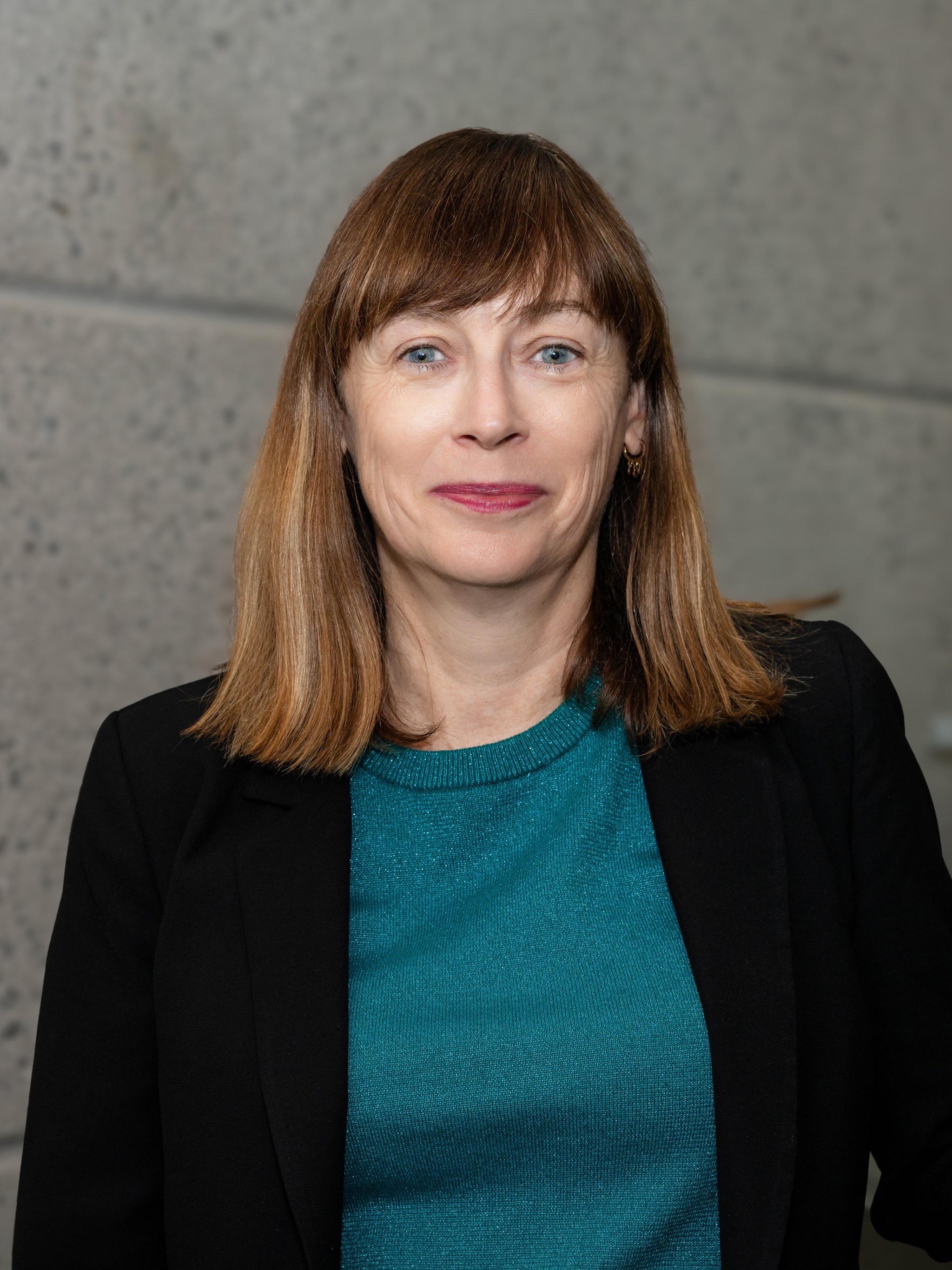 A woman with shoulder length brown hair, wearing a dark blazer over a blue-green top.