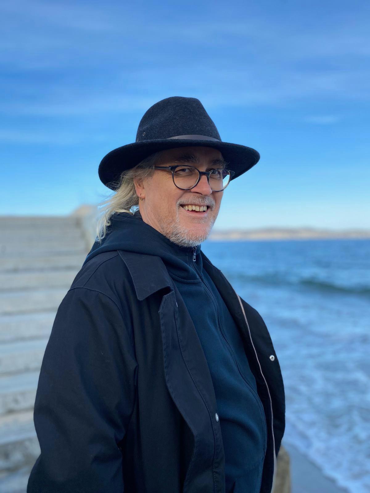 A mid-shot of man (Tim Sherratt) with long grey hair and a short grey beard wearing black glasses and a black wide-brimmed hat smiling. Behind him are concrete stairs and a beach shoreline.