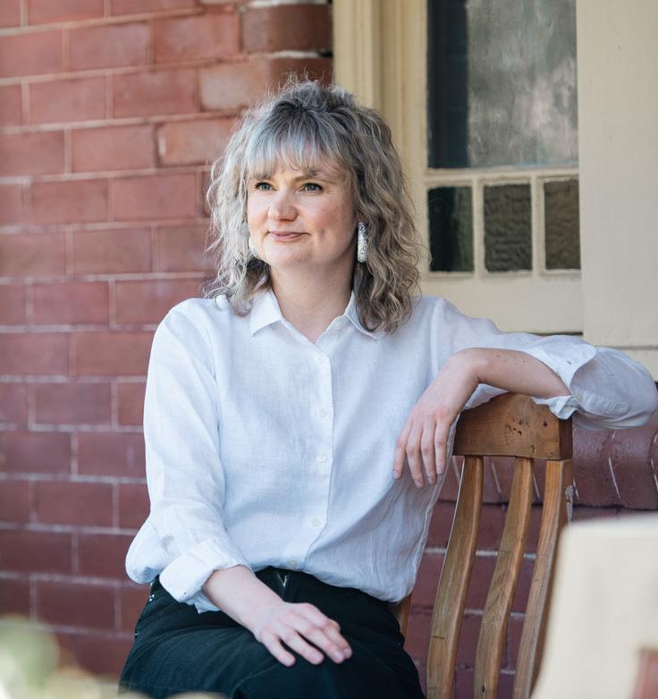 Portrait of woman (Vanessa Bartlett) with light shoulder length hair. She is sitting in a wooden chair outside and looking out to the left of shot. She is wearing a white shirt.