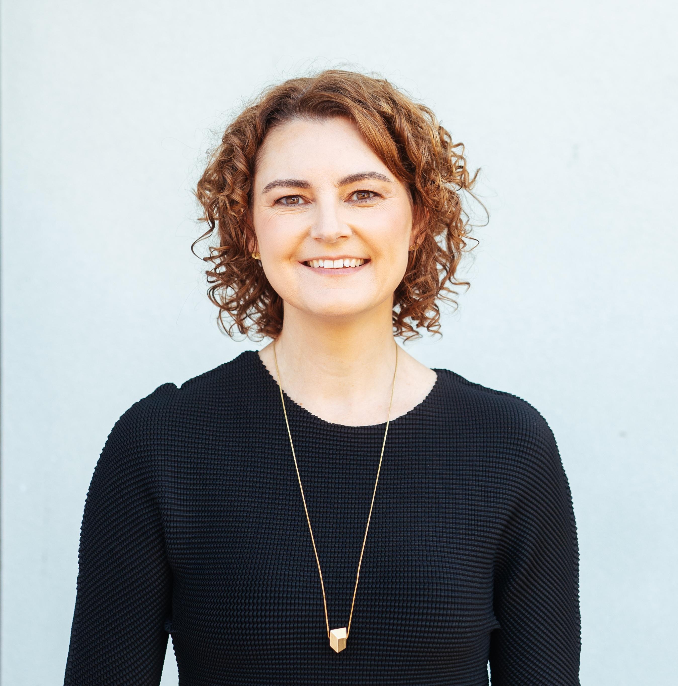 A portrait front-on shot of a smiling woman (Angela Stengel) with medium length curly auburn hair wearing a black top and gold necklace