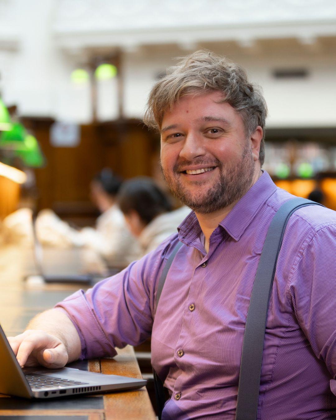 A portrait of smiling man (Ben Kolaitis) with short brown hair and a light beard wearing a purple shirt and grey suspenders seated at a desk.