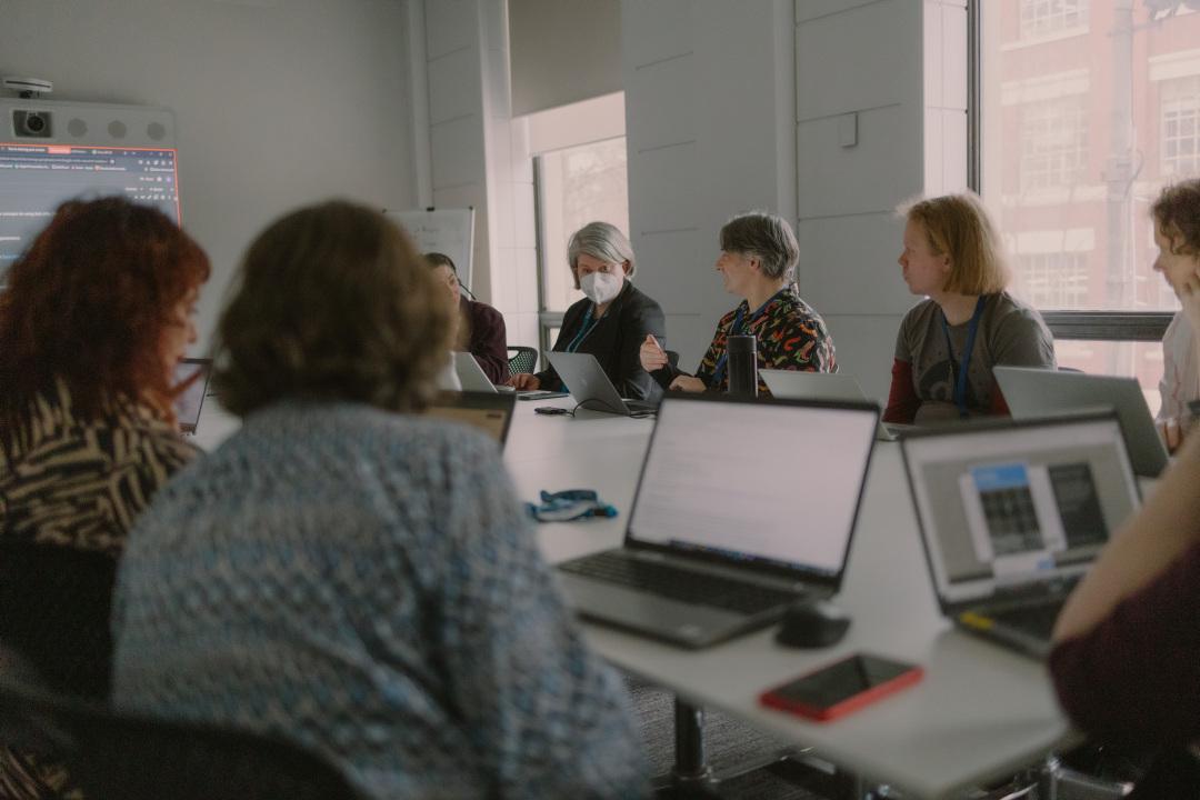 Group of people sat around a desk having a discussion. They are in a meeting room with laptops in front of them.