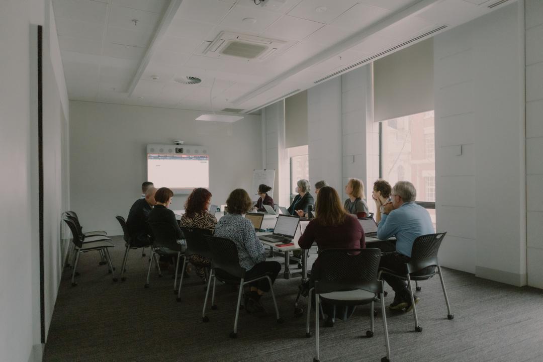 A group of people sitting around a desk in a meeting room, each using a laptop. There is a large screen at the front of the meeting room.