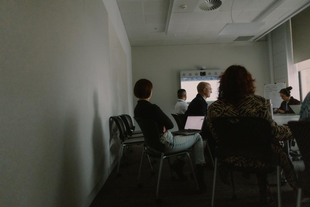 A small group of people in a meeting room, one of whom is sitting back in their chair. There is a large screen at the front of the room.