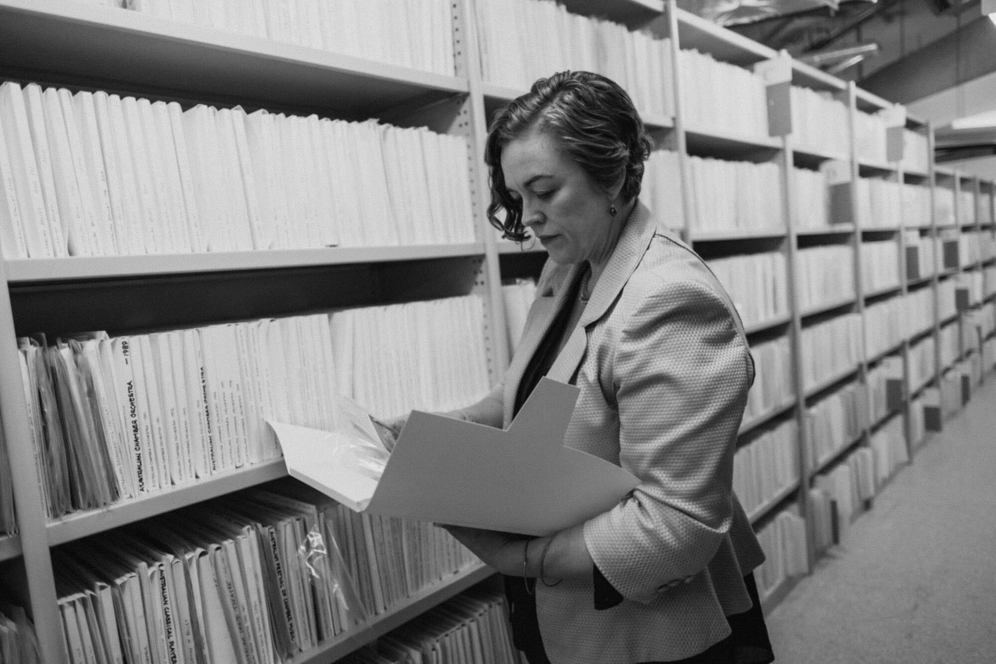 Person wearing a blazer examines a folder in an archive room filled with shelves of files.