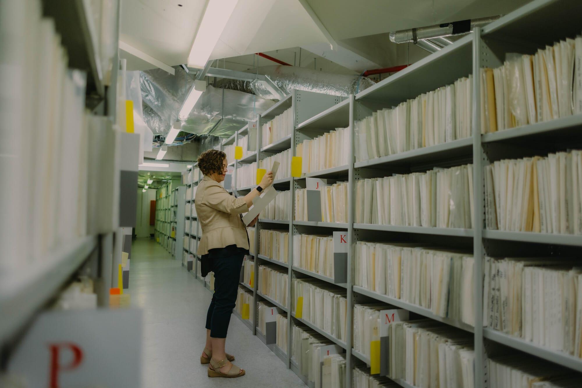 Person browsing rows of archived files in a large, well-lit storage room filled with shelves labeled alphabetically.