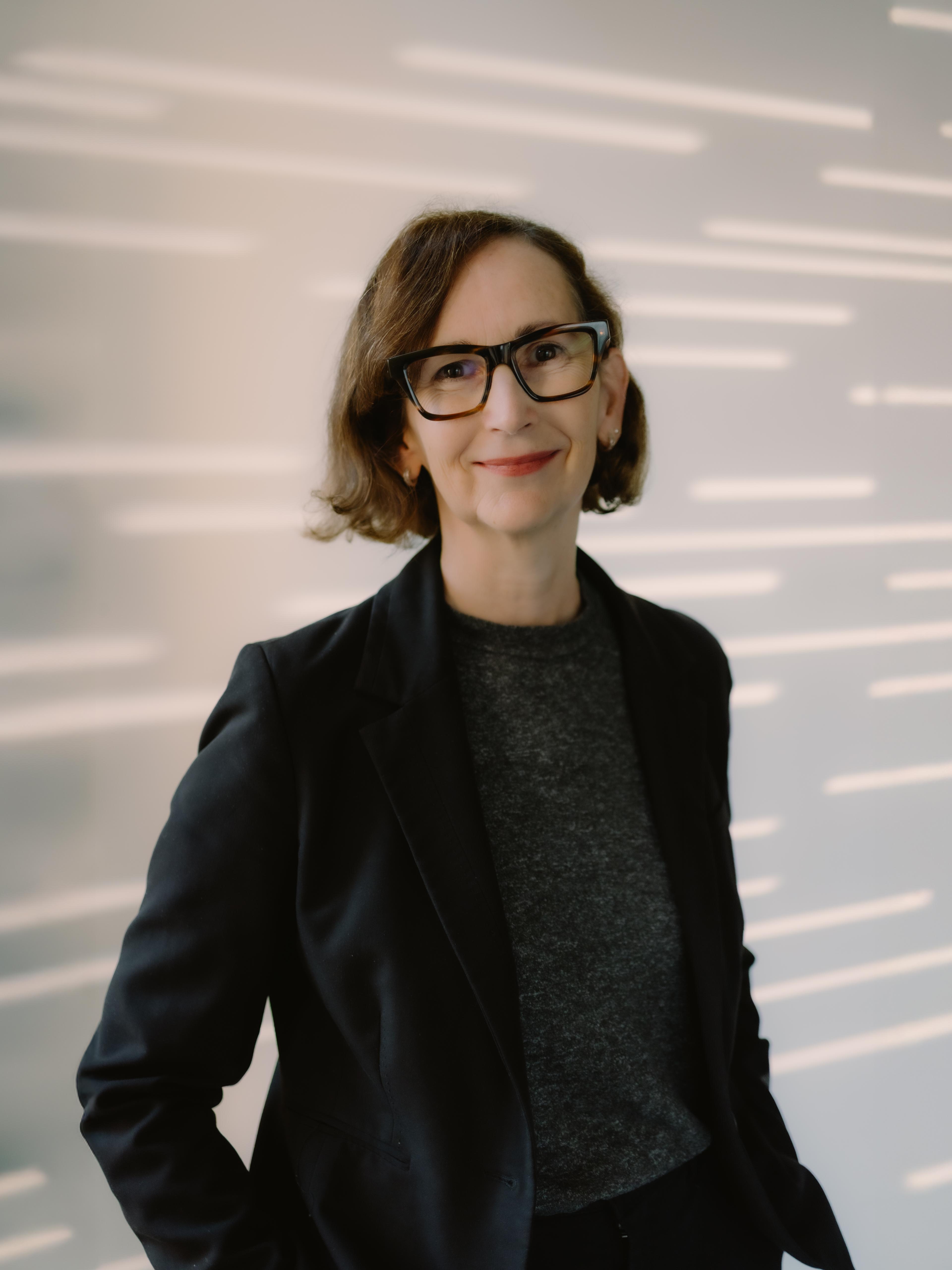 Headshot of a smiling woman with short brown hair, wearing a blazer and dark glasses, standing in front of a decorative wall.