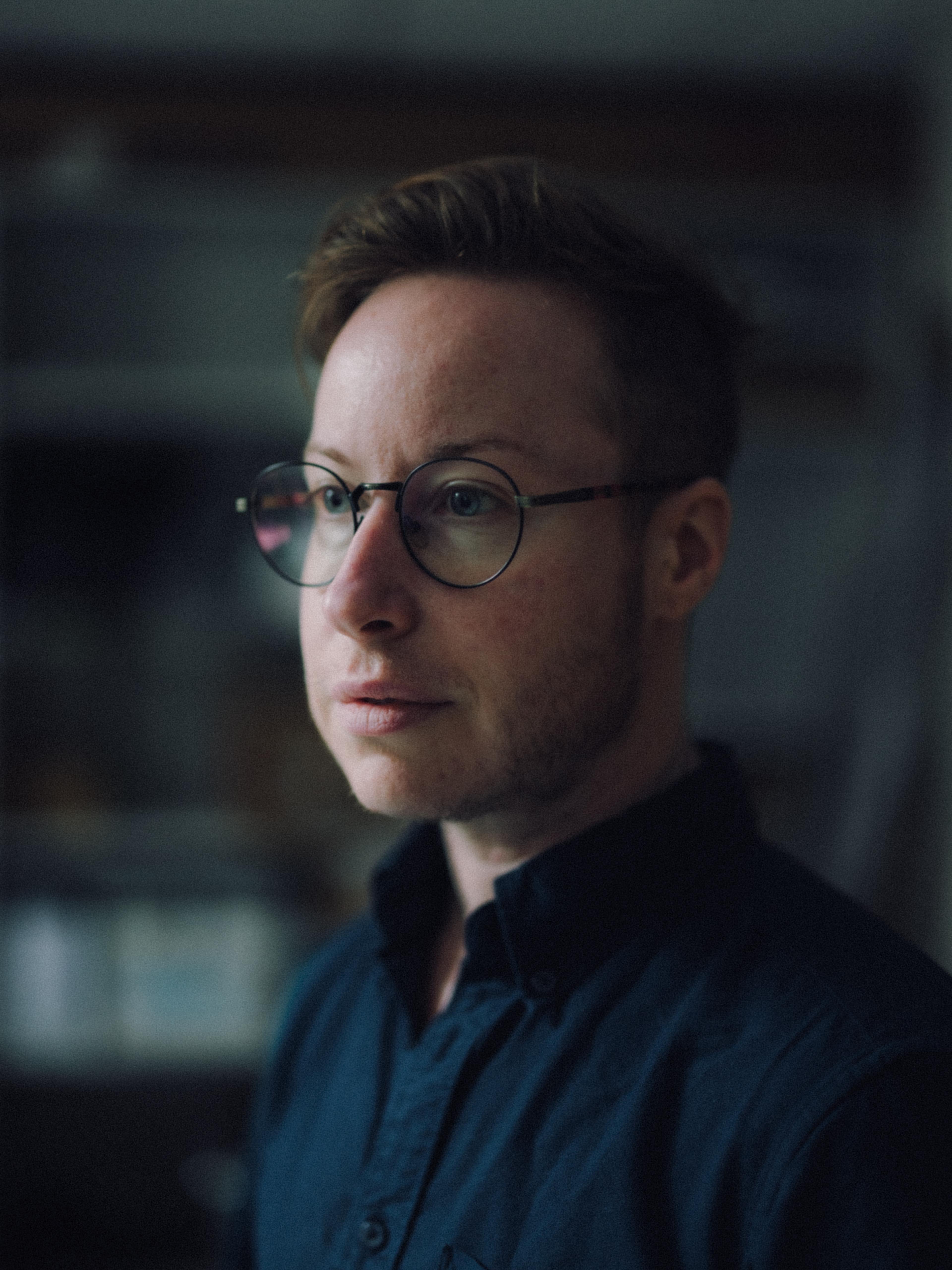 Headshot of a bearded person, wearing rounded glasses and looking off to the side.