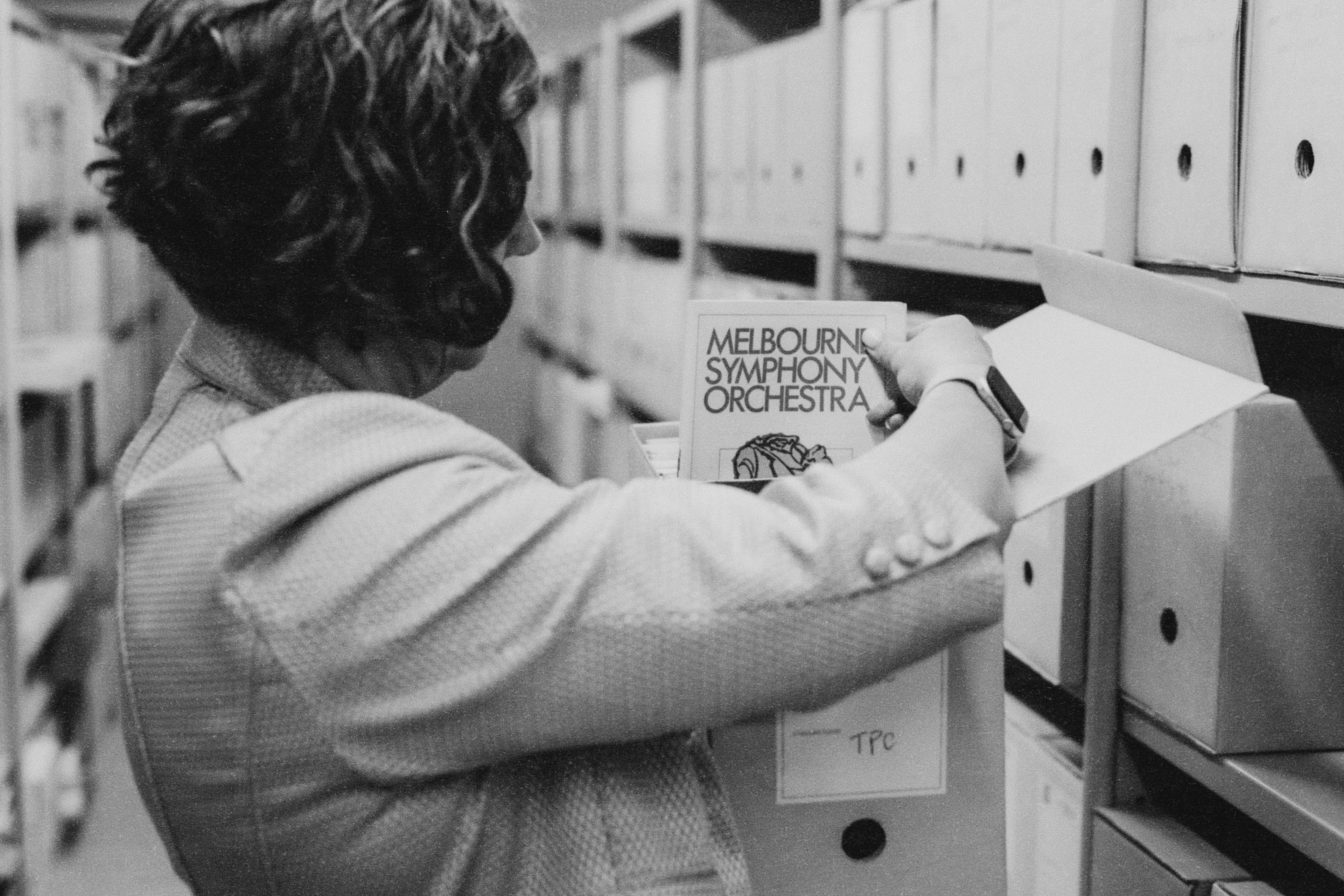 Person holding an archival box, looking at a Melbourne Symphony Orchestra program in front of a shelf full of archival boxes.
