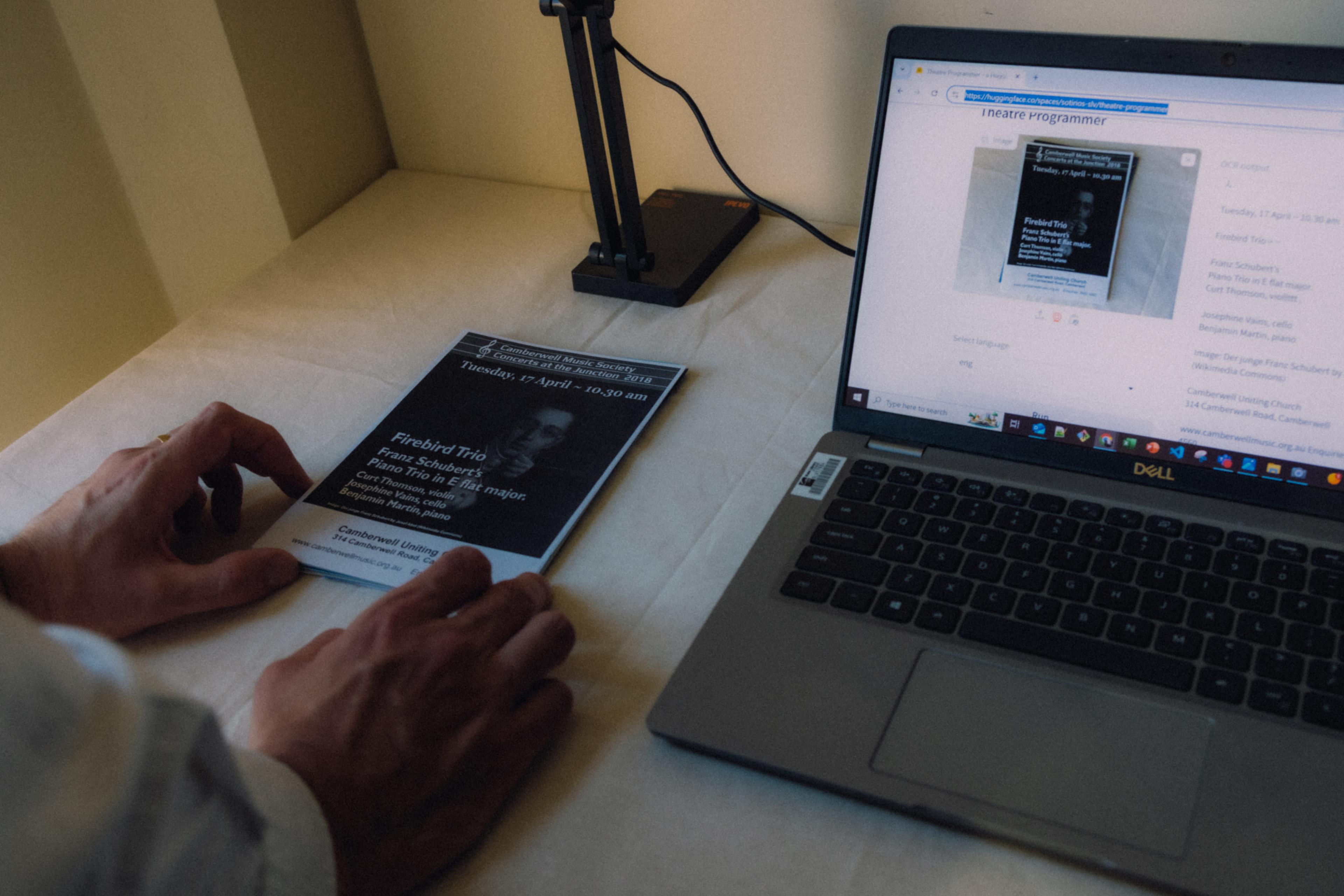 A pair of hands adjusting a theatre program under a device sitting next to a laptop on a desk.