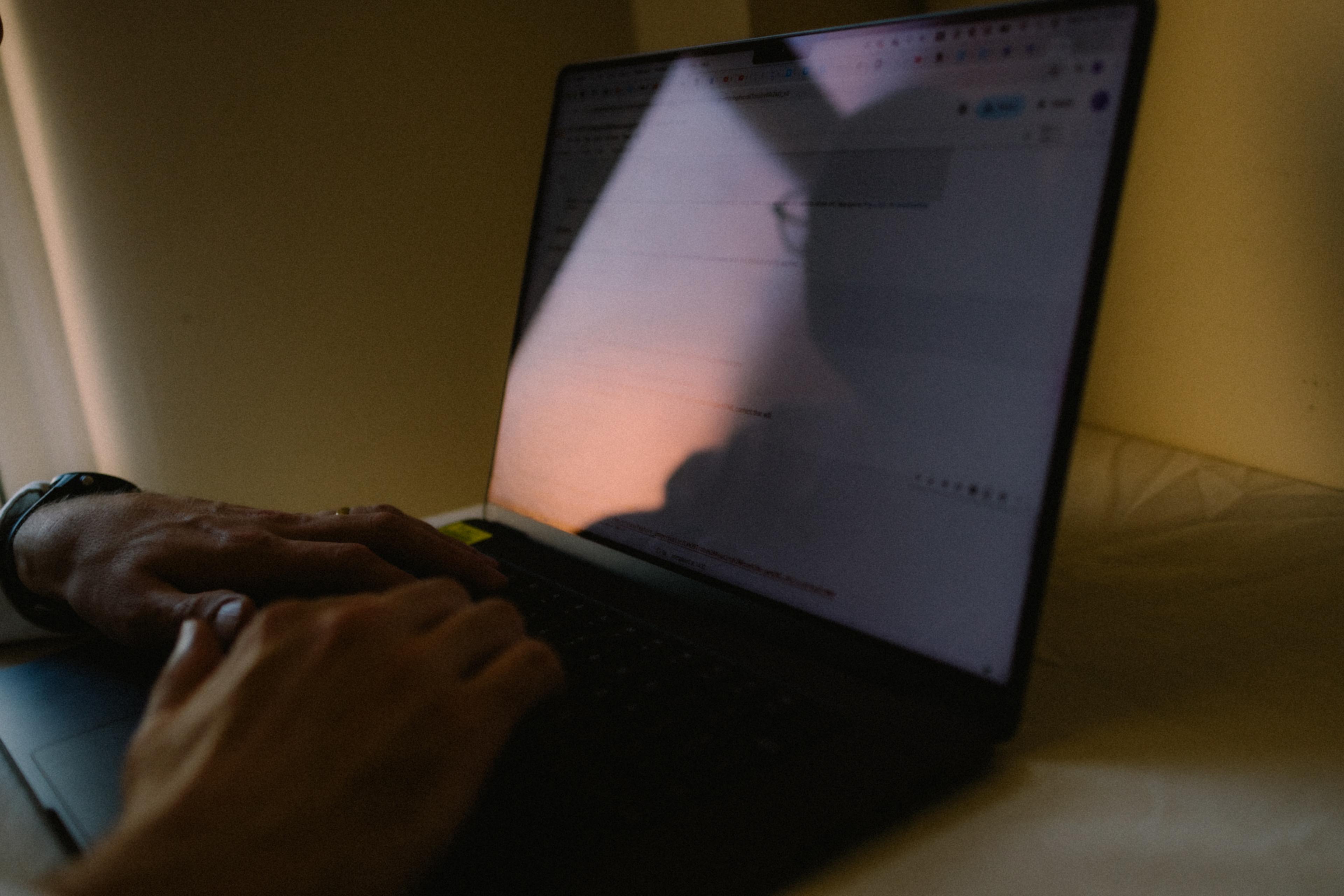 A laptop with a pair of hands on the keyboard. A person's face is partially reflected in the screen.