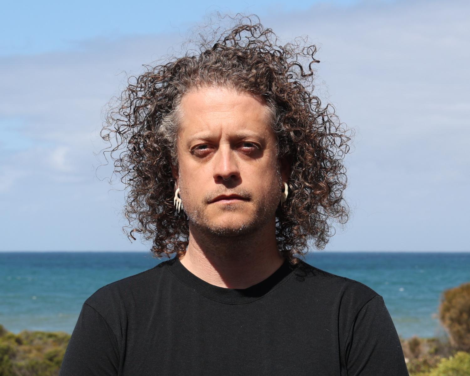 Headshot of a serious-looking person with long curly hair, wearing cream-colored abstract earrings and standing in front of a large body of water.