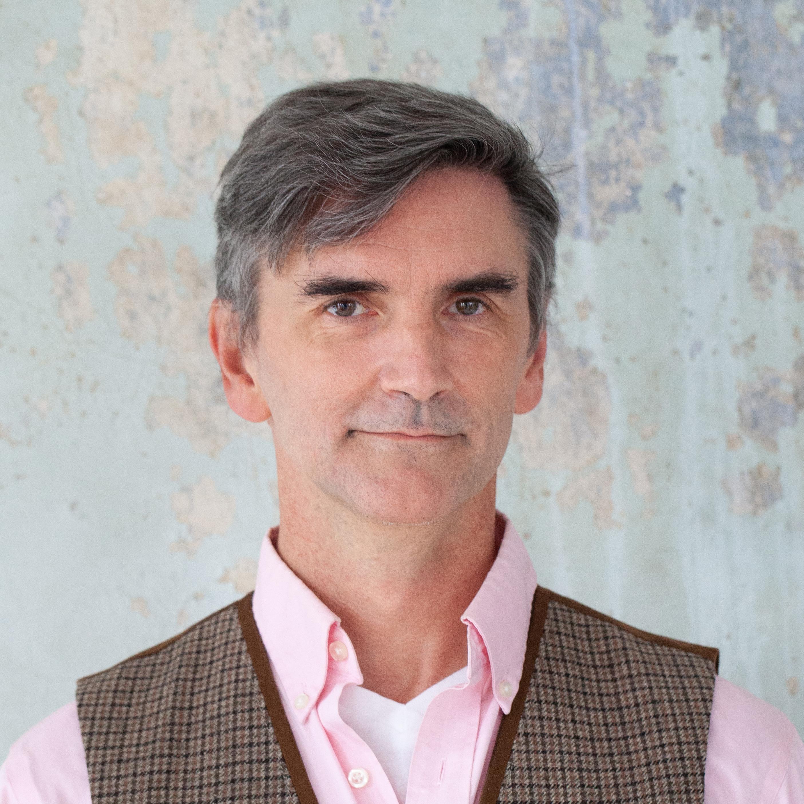 Headshot of a man with dark, greying hair. He is wearing a pale pink shirt and a checked waistcoat.