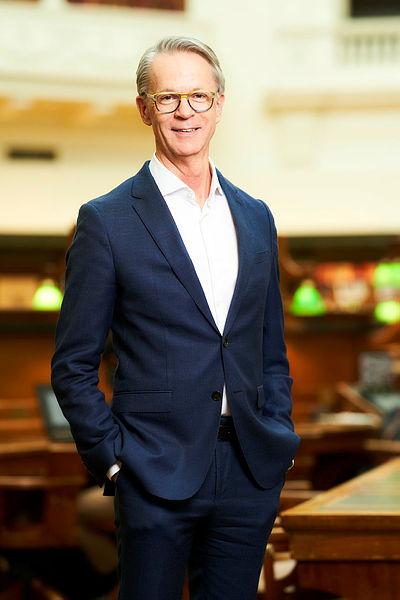A smiling man wearing a blue suit and glasses, standing in front of blurred library desks and interior architecture.