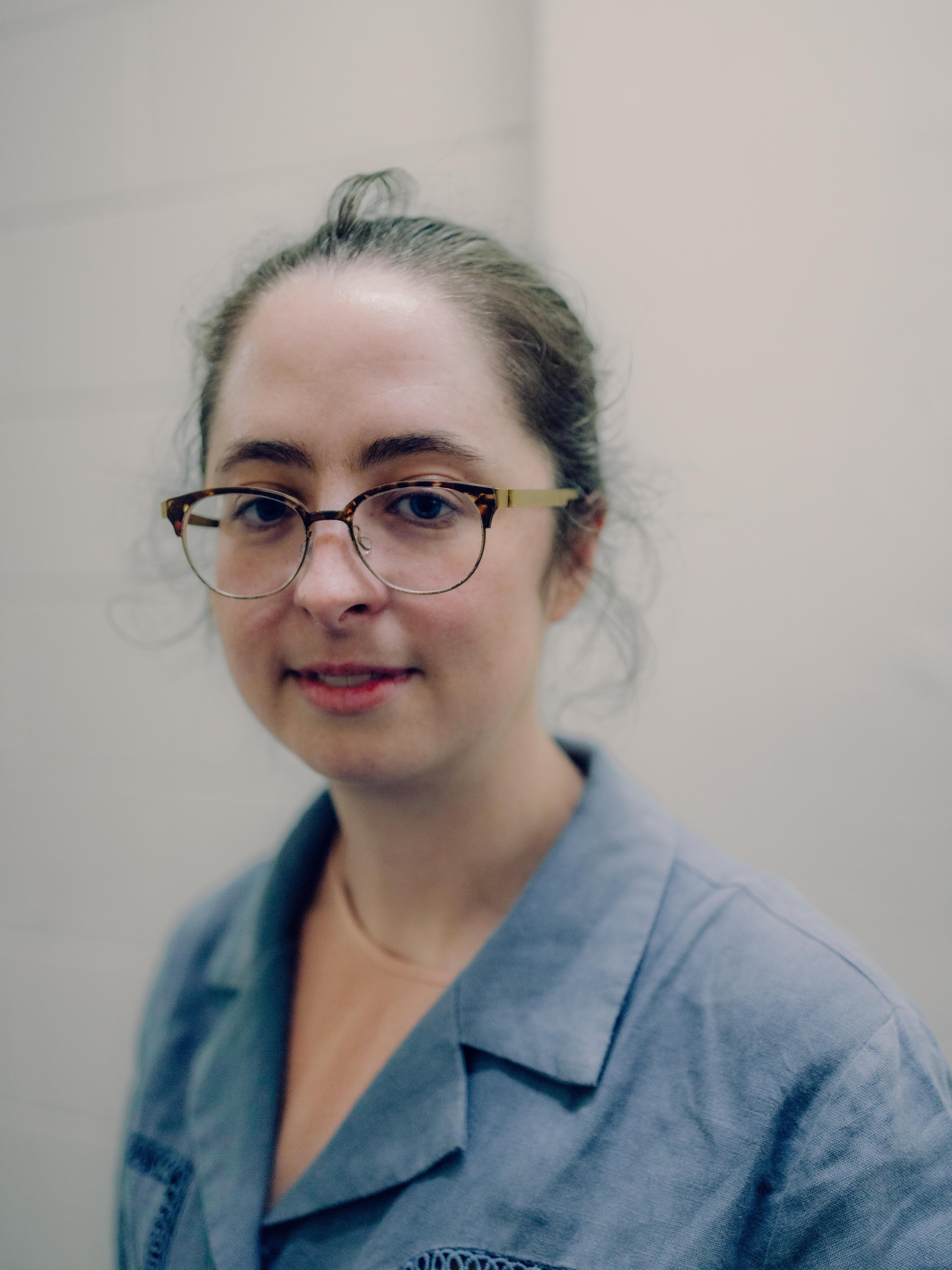 Headshot of a bespectacled woman with dark hair, wearing a blue collared shirt.