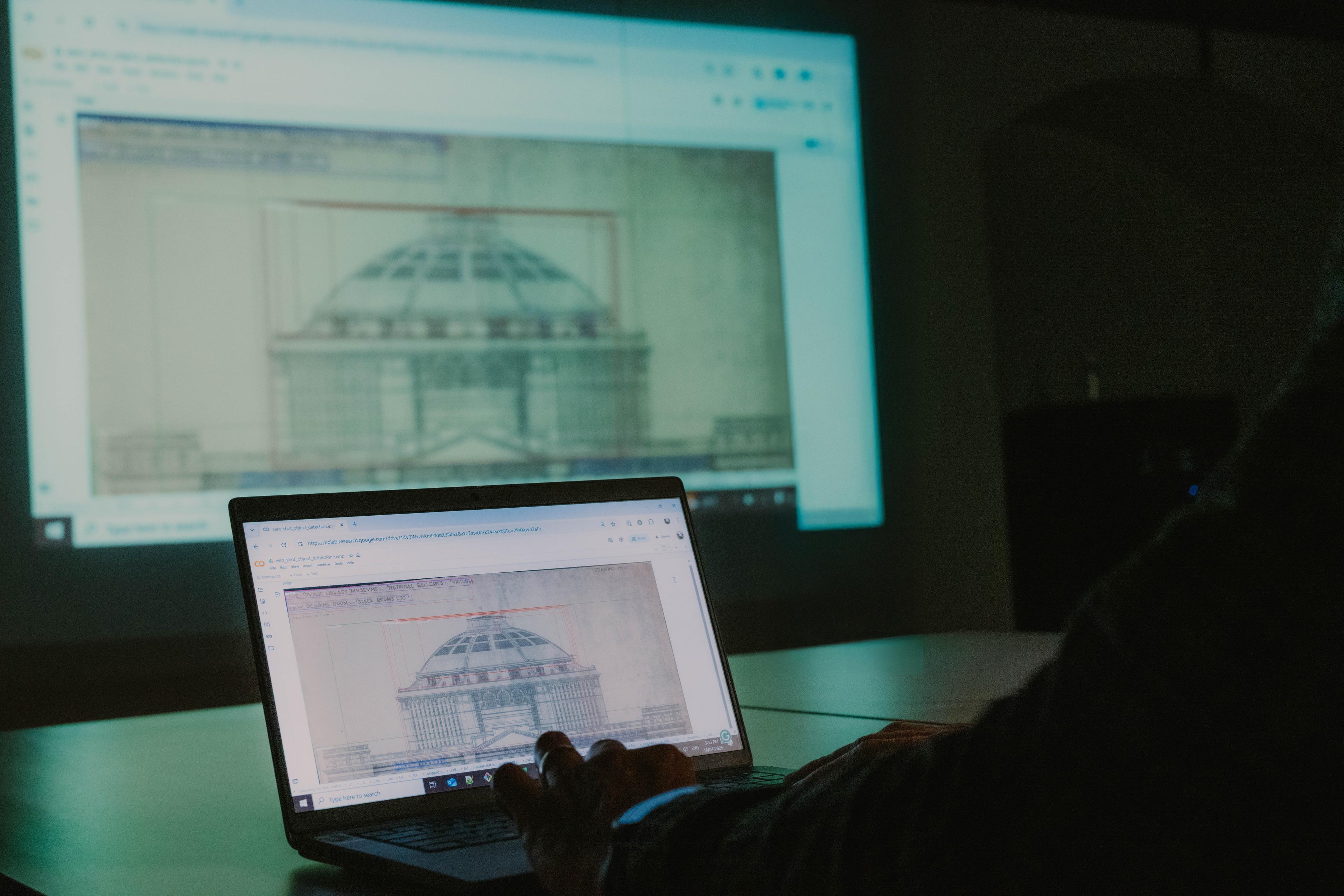 A hand typing on a laptop that displays an architectural drawing. The display is mirrored on a large screen in the background.