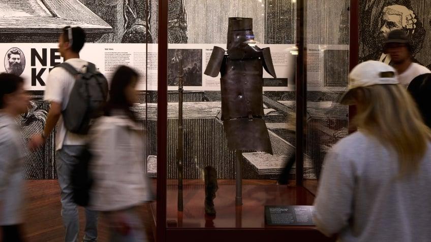 Rusted armour suspended in a glass case, surrounded by blurred people walking past, set against a wall of explanatory images and text.