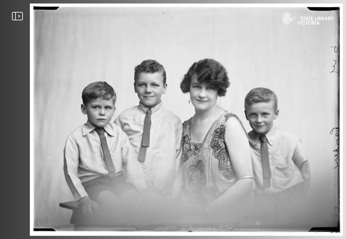 Black-and-white photograph of three boys and a woman. Each are facing the camera and are dressed in formal clothing.