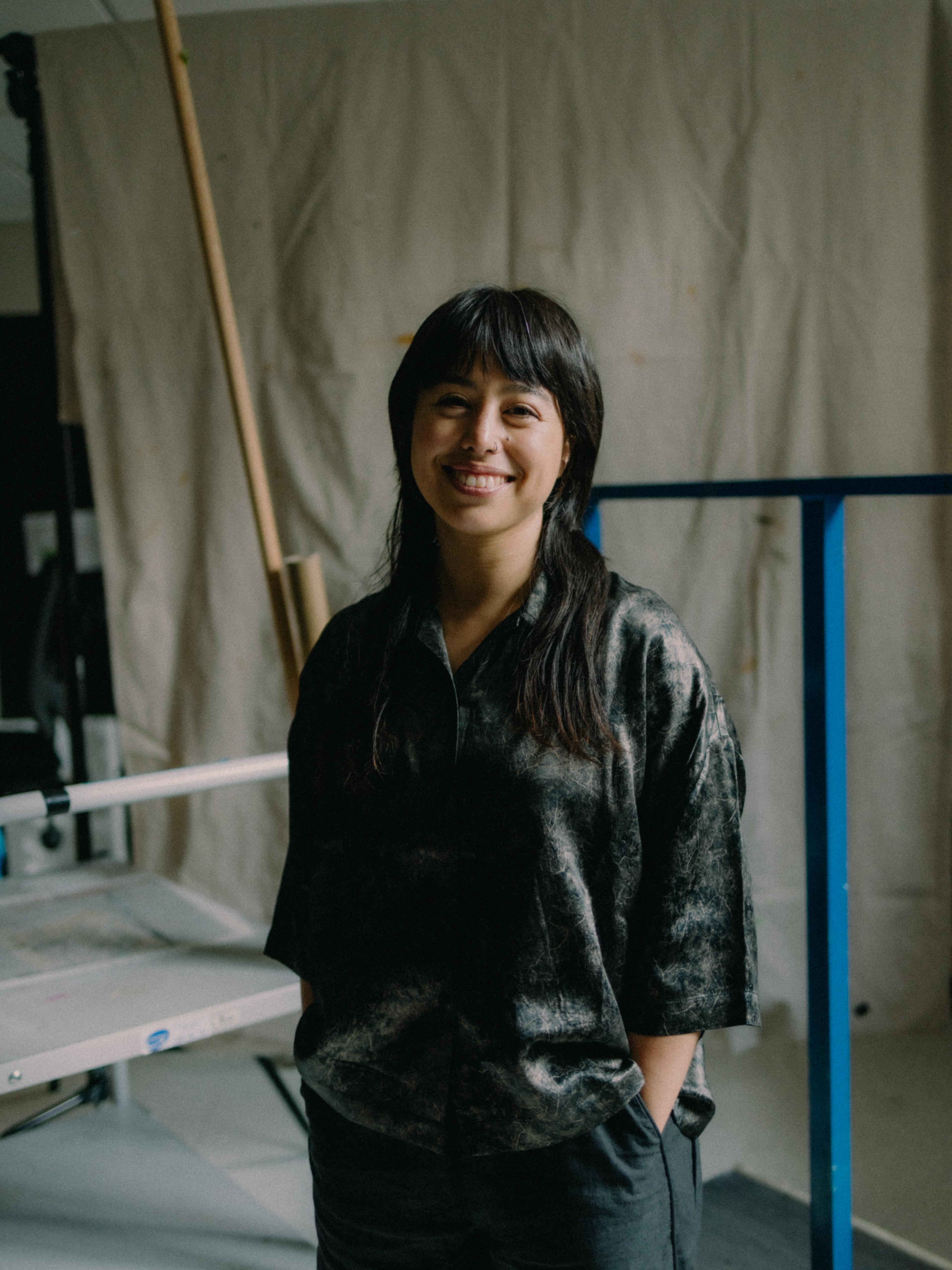 A person with dark hair and a dark, petrol blue shirt stands and smiles at the camera.