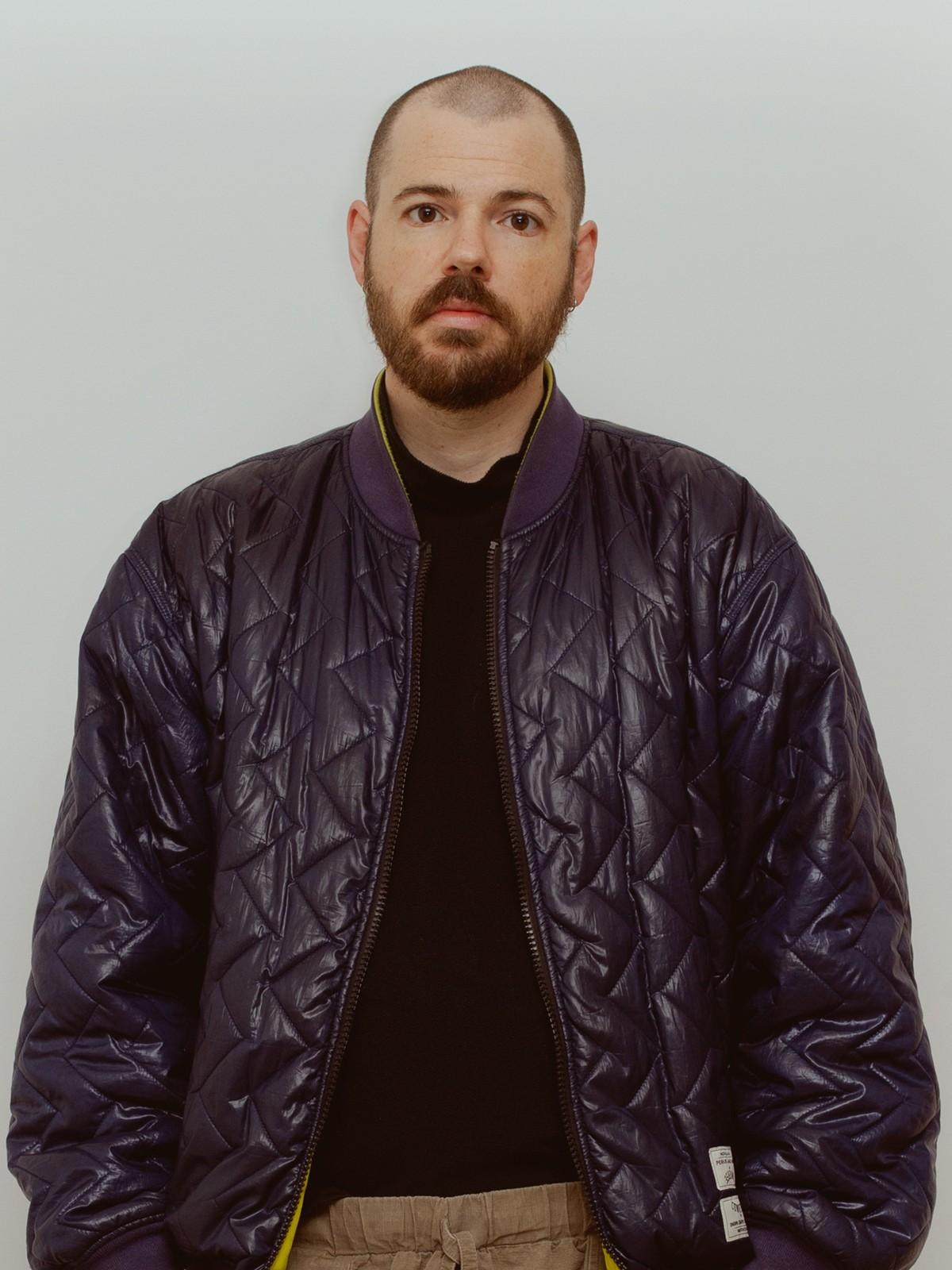 A bearded man wearing a purple puffer-style bomber jacket over a black shirt, looking straight at the viewer with a neutral expression.
