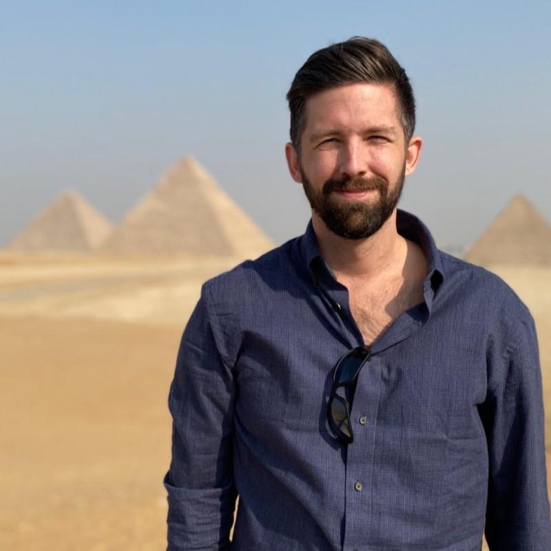 A bearded man, in a dark blue shirt, squints in the sun. In the background is a desert and pyramids.