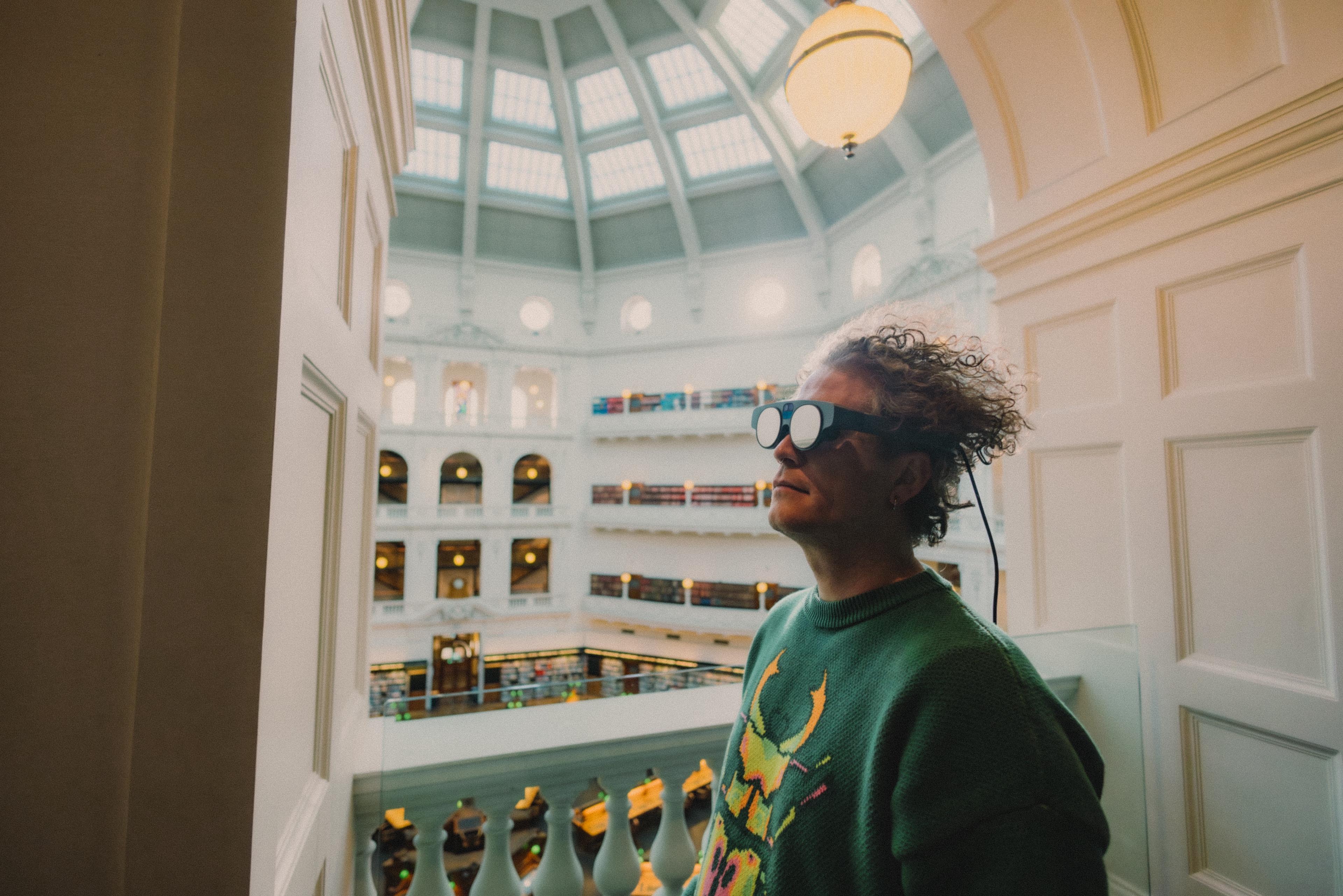 A person in a green jumper, with curly hair wearing reflective glasses looks off to the left in front of a large room with a domed ceiling. 