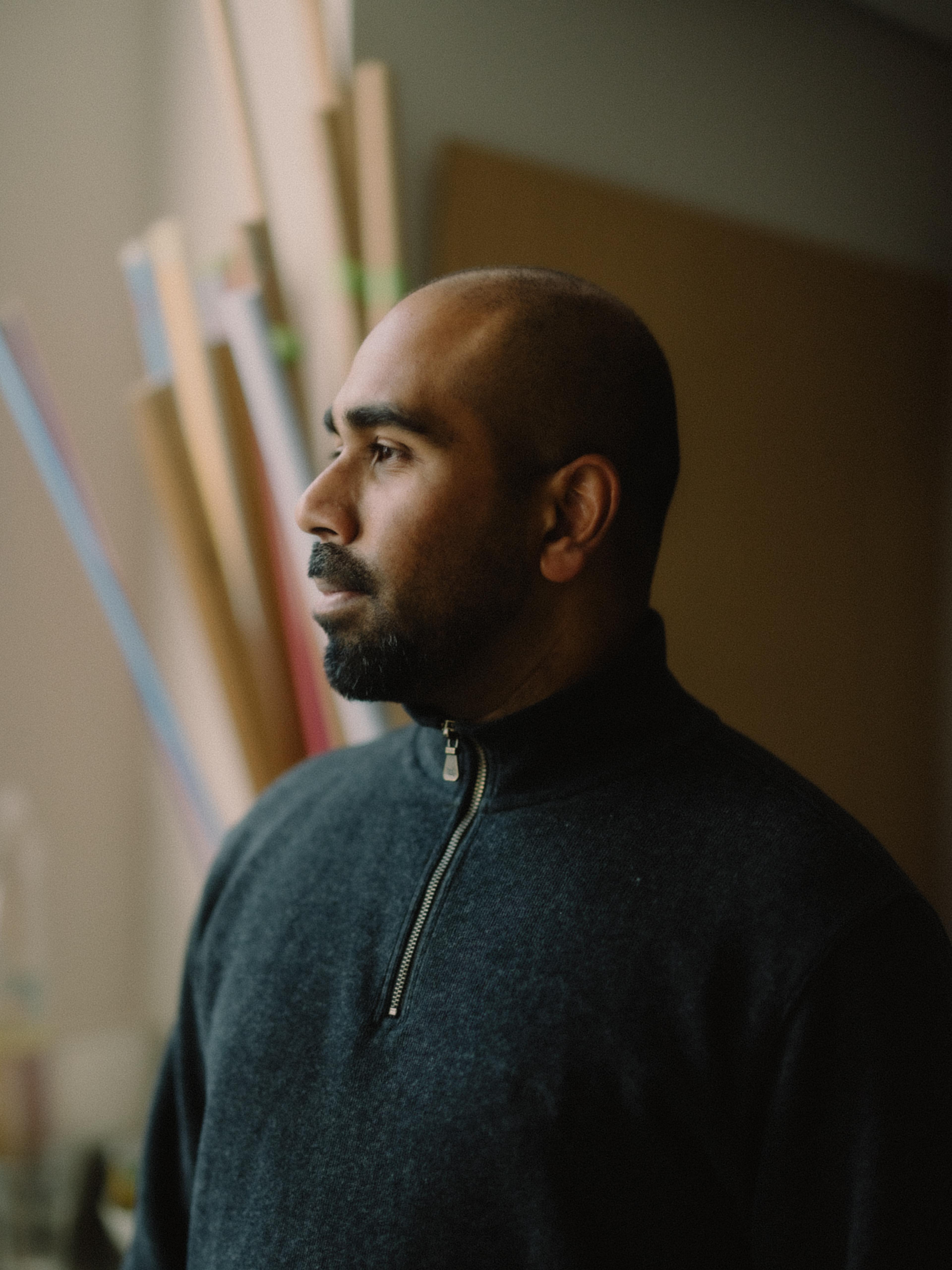 A side profile of a dark-skinned bald man with a beard wearing a dark blue cardigan