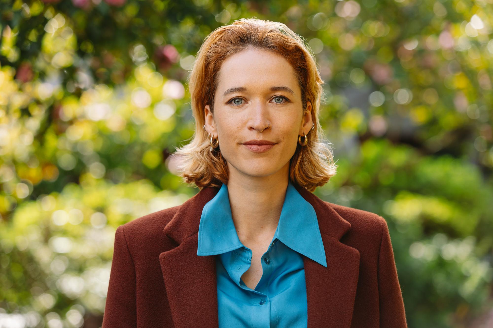 A headshot of a woman (Jasmin Pfefferkorn) with shoulder length brown hair, blue eyes. She is wearing a bright blue shirt and a maroon blazer.
