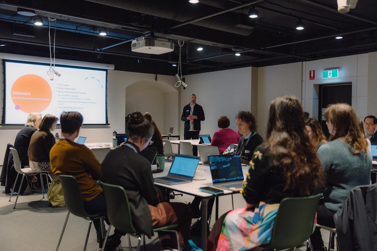 A group of people seated at tables with laptops in front of then. They are facing a speaker (Sotirios Alpanis). The event is a workshop called WikiFest 2025