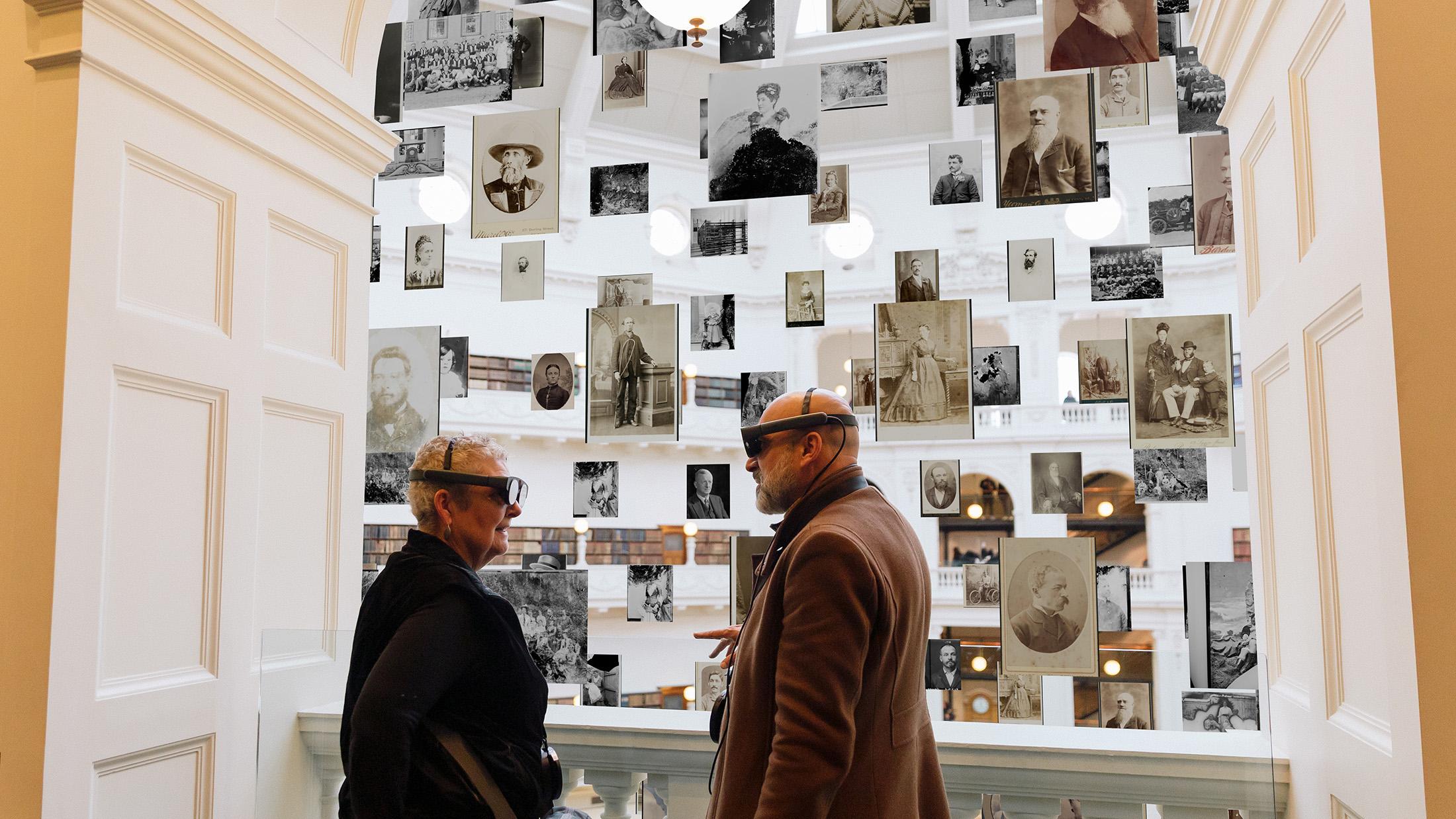 Two older people wearing mixed reality headsets talking to each other as black and white and sepia images float around in a large, multi-storey gallery space behind them