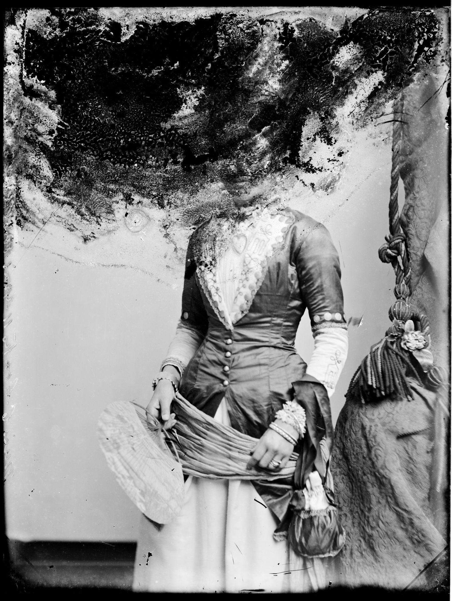  A black and white photograph of a Victorian-era woman in an elegant dress posing for a studio portrait. Her head has been completely obscured by damage to the photographh
