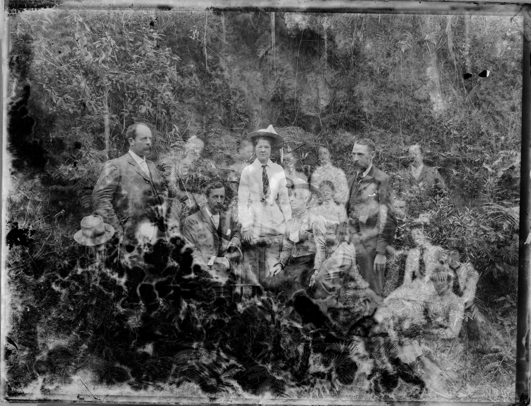 A black and white photograph of a group of men and women in Victorian era attire posing in a bush setting. The photograph is heavily deteriorated with ghostly double exposures and burn marks