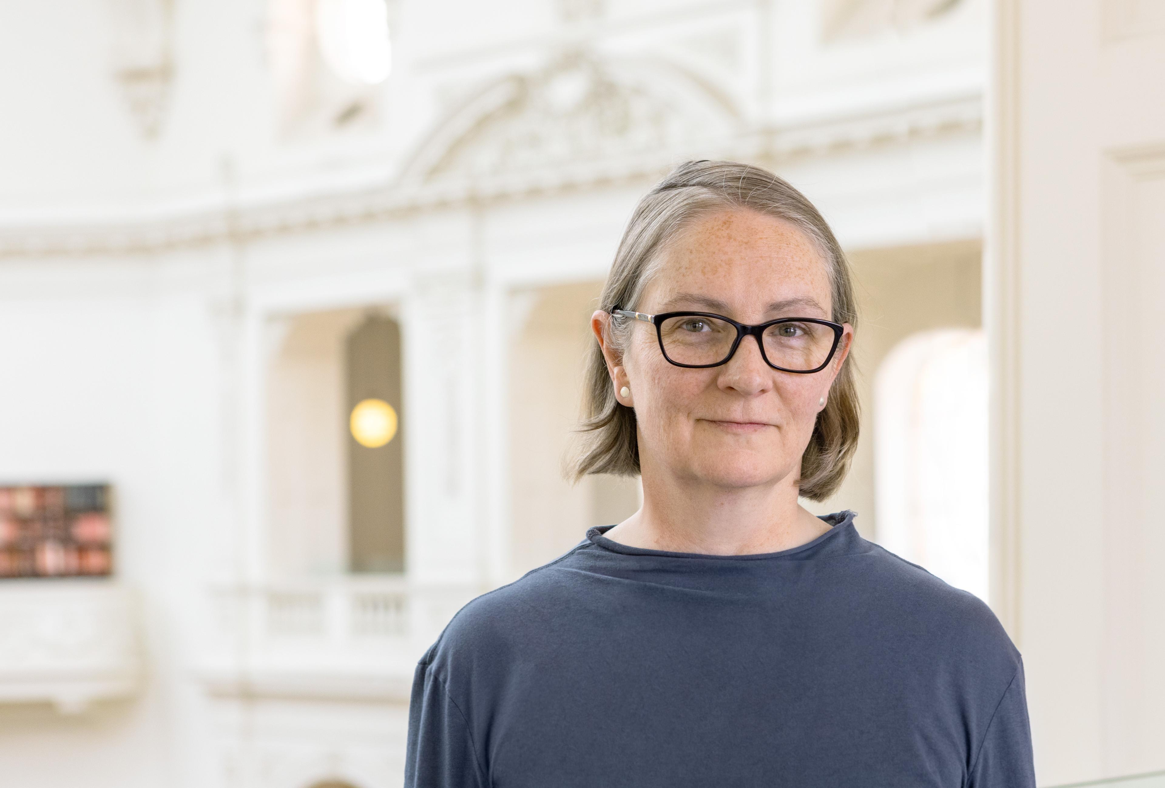 A woman (Vanessa Ross) with short grey-brown hair wearing a blue top and black glasses standing in an ornate white library space