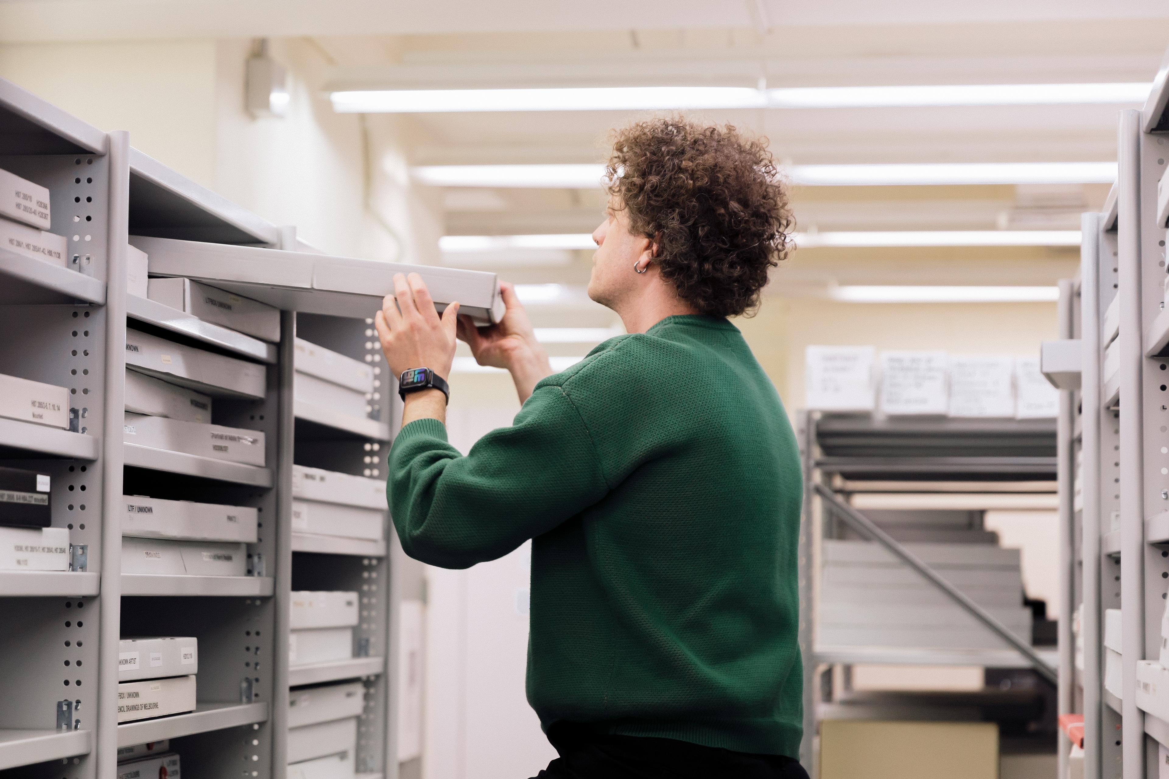 A person (Yoyo Munk) wearing a green sweater looking through boxed archival records on shelves. Their back is to us and they appear to be standing on a stepladder
