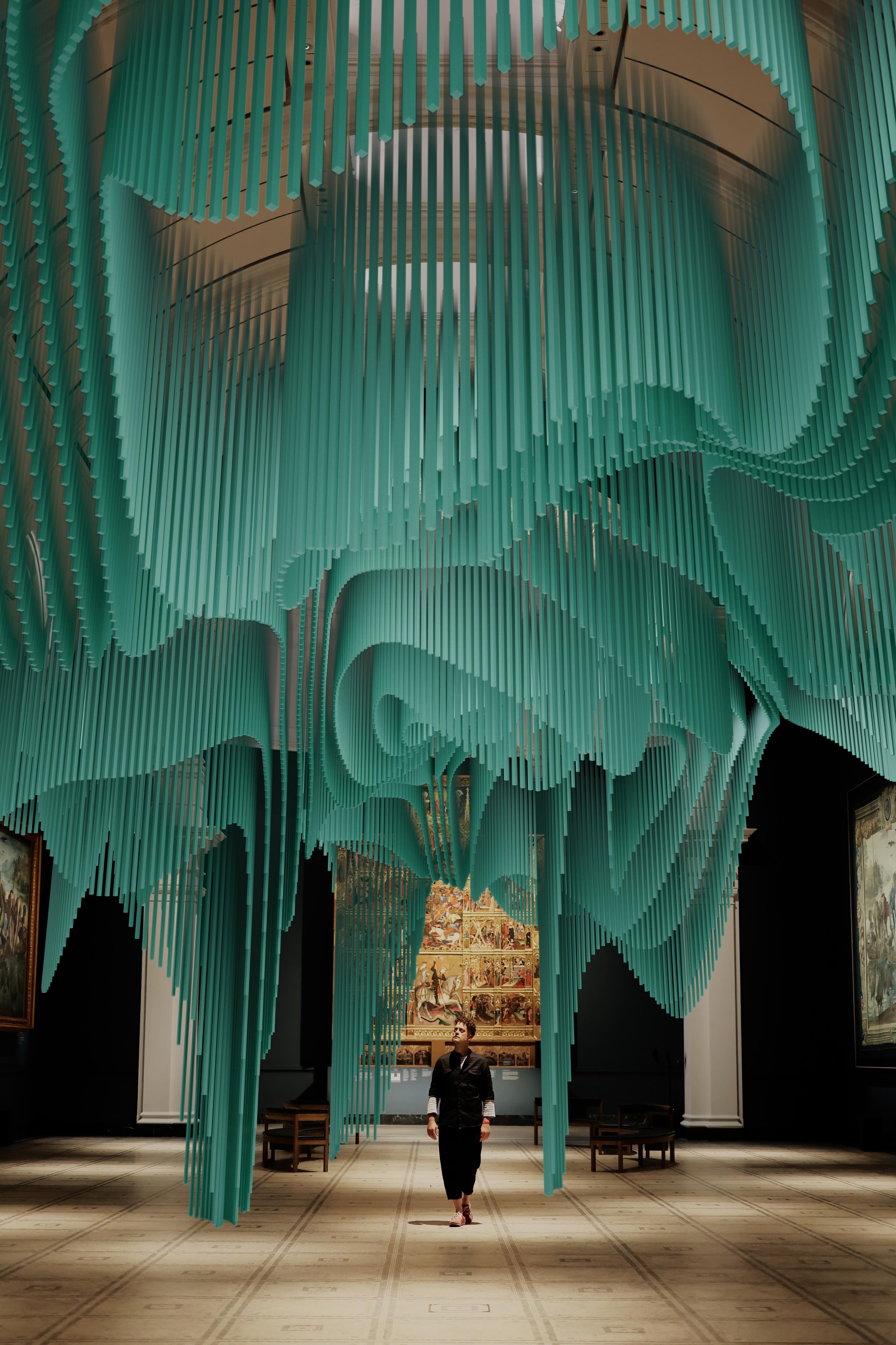 A large scale digital installation of many green-blue columns forming wave-like patterns on the roof space of a gallery. A person (Yoyo Munk) is walking underneath it