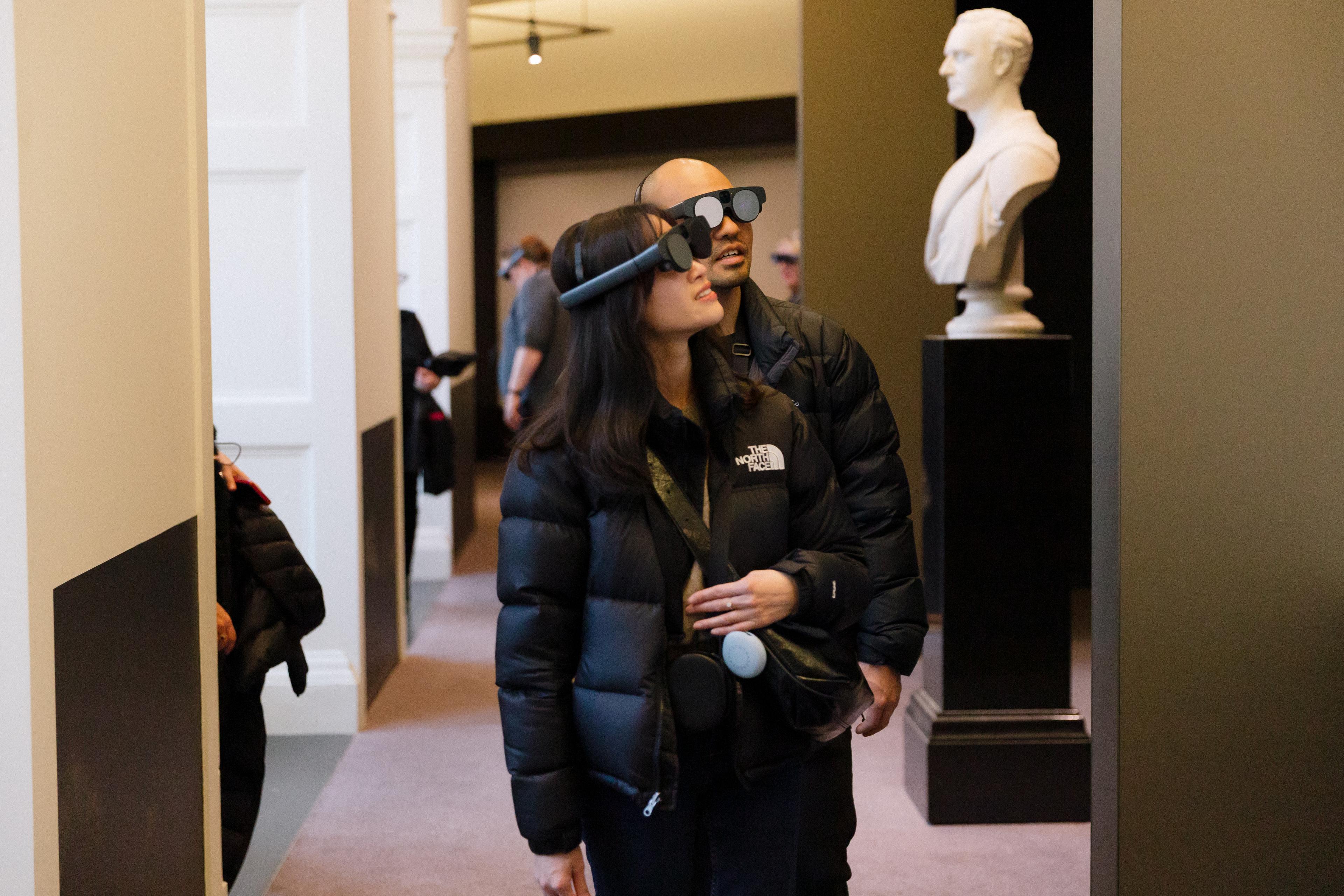 Two people of asian appearance wearing mixed reality headsets and looking up at the walls of the gallery space. There is a white marble bust of Redmond Barry behind them