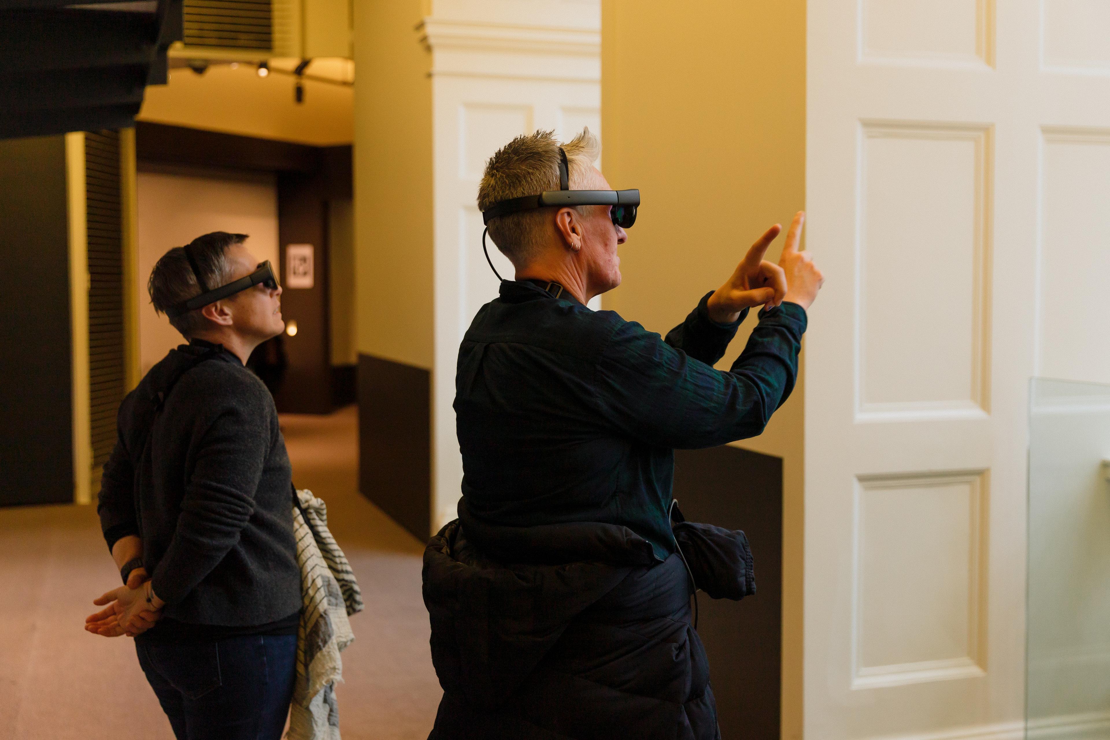Two people wearing mixed reality headsets, the person in the foreground is marking a framing gesture with his fingers