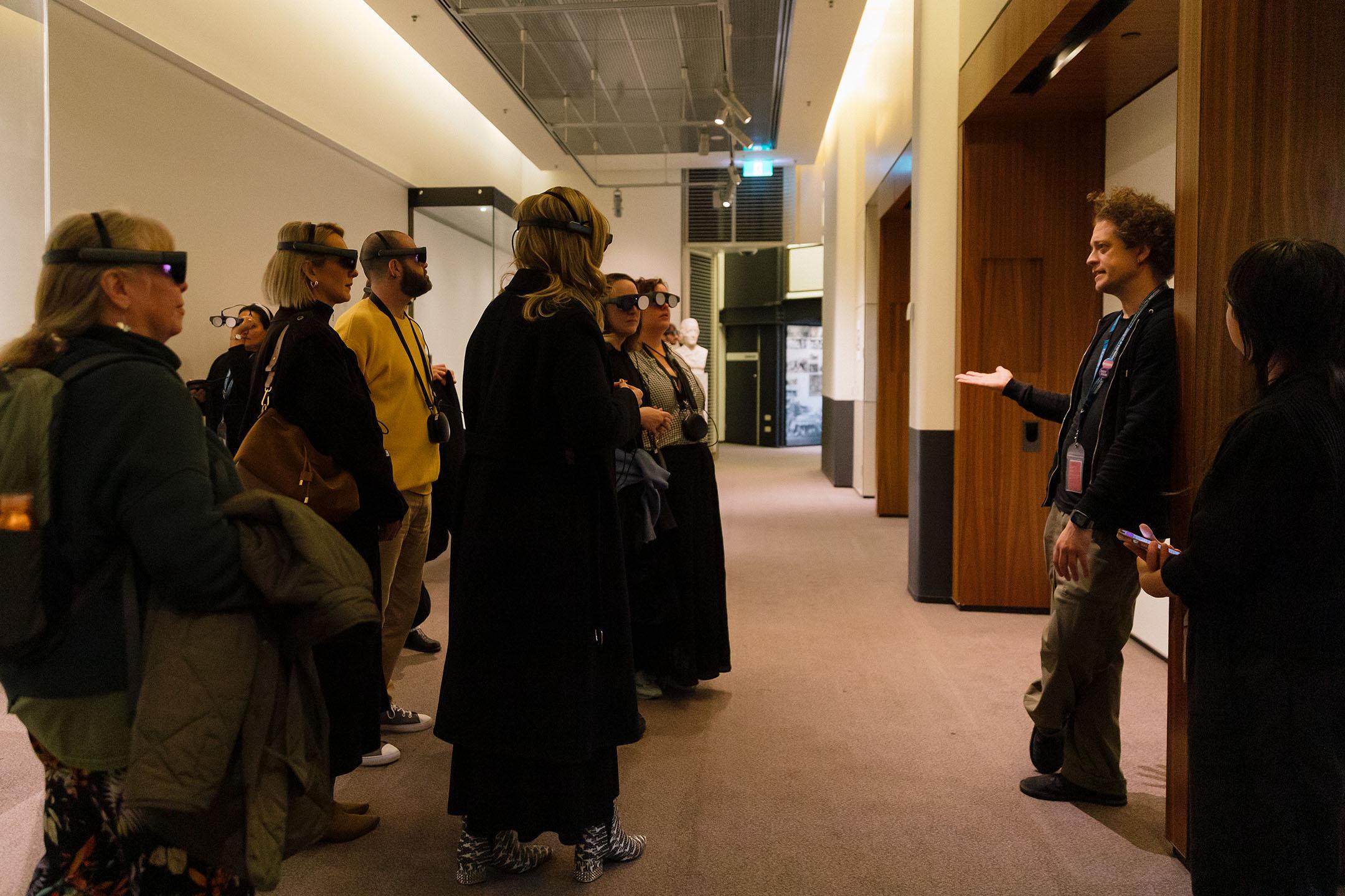 A small crowd wearing mixed reality headsets listen intently to an introductory speech by artist Yoyo Munk