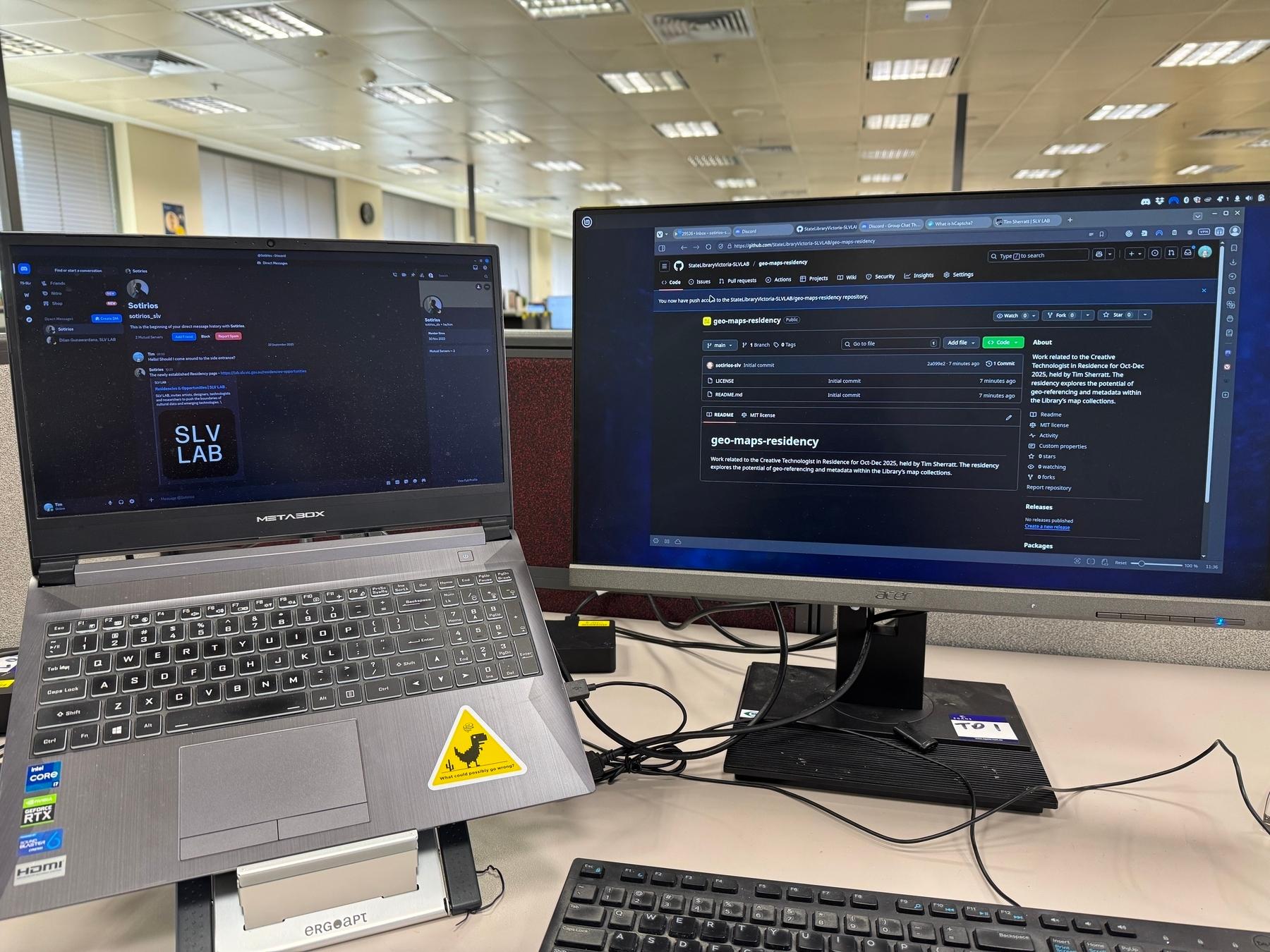 A laptop and a desktop screen side by side in an office - the laptop shows the SLV LAB logo on it, while the desktop displays a github repository