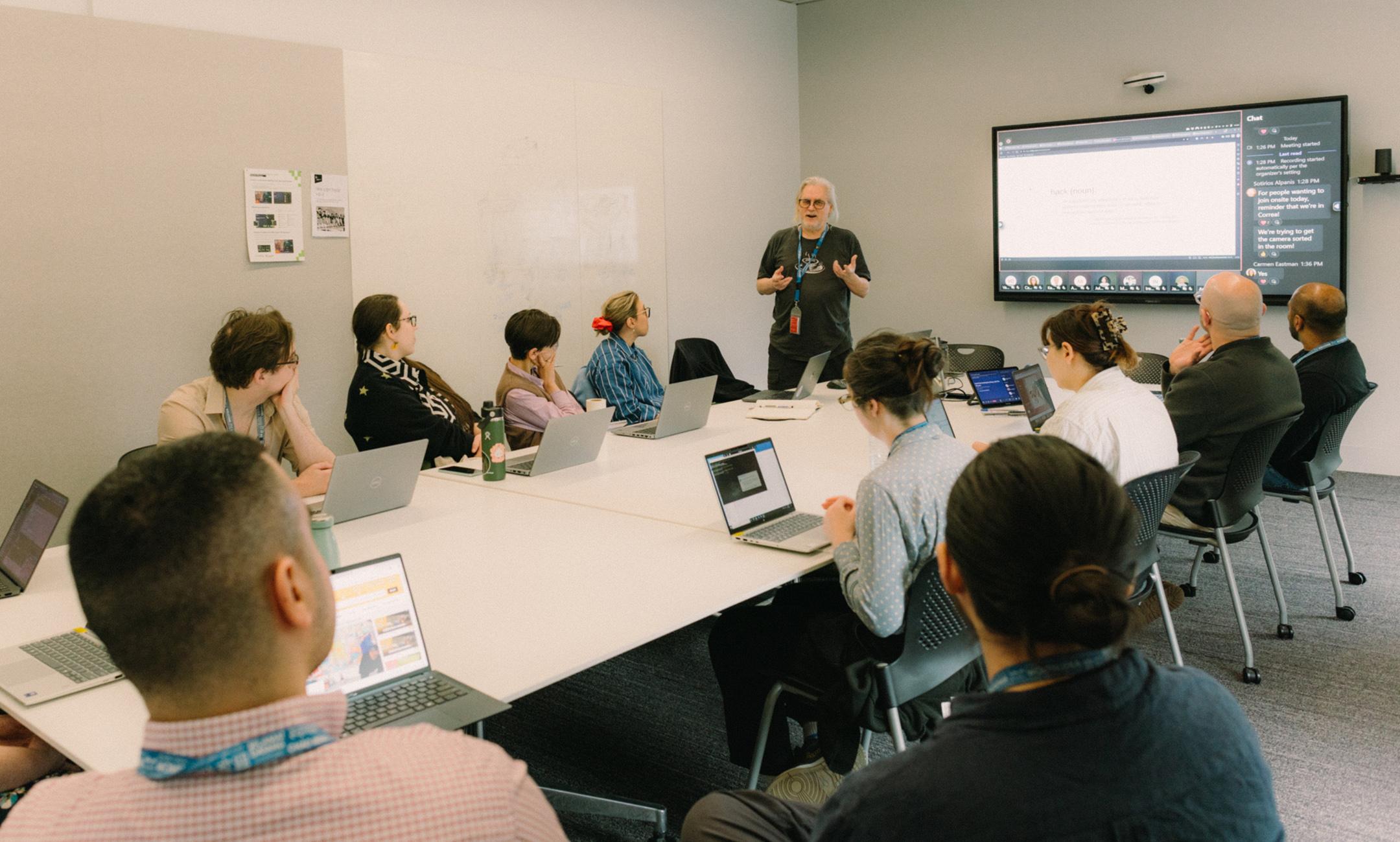 A man with long white hair wearing black glasses and a tshirt (Tim Sherrat) gesturing as he presents to a room of people in front of their laptops