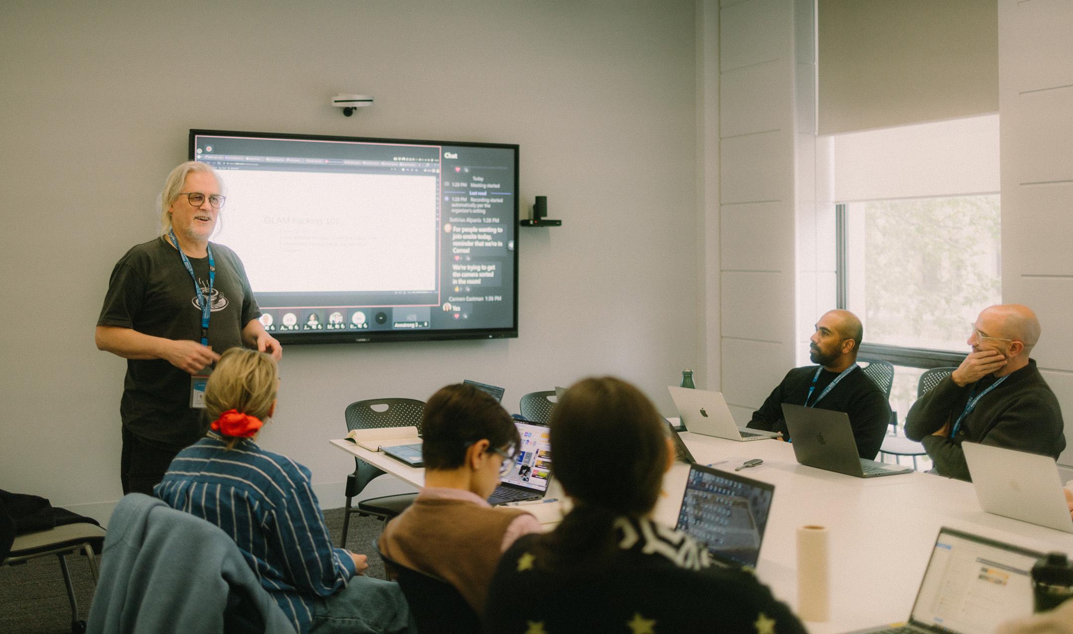 A smiling man with long white hair wearing black glasses and a tshirt (Tim Sherratt) speaking to staff at a workshop