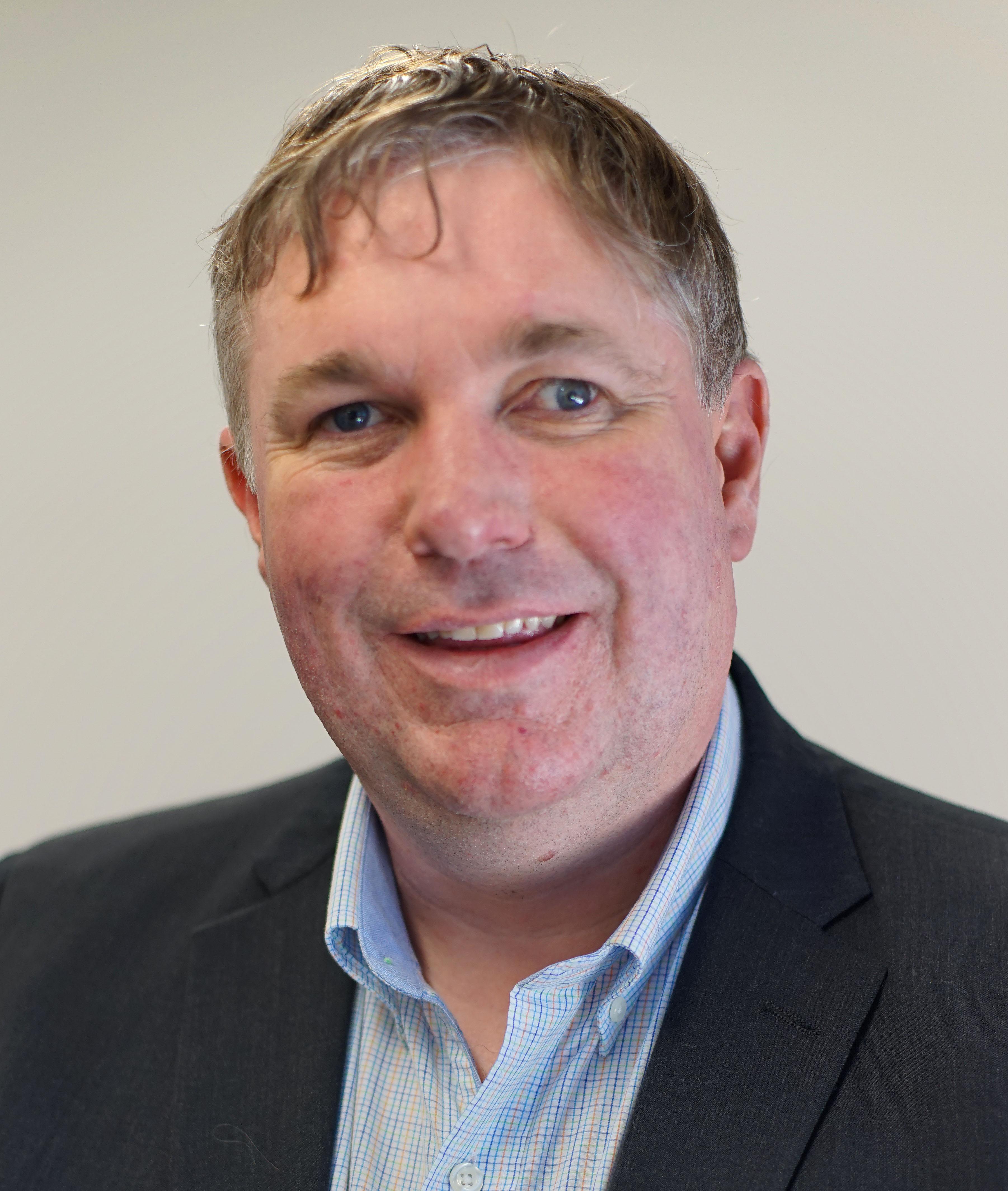 A closeup portrait of a smiling man with short brown-grey hair wearing a black blazer and blue shirt - Scott Hollier