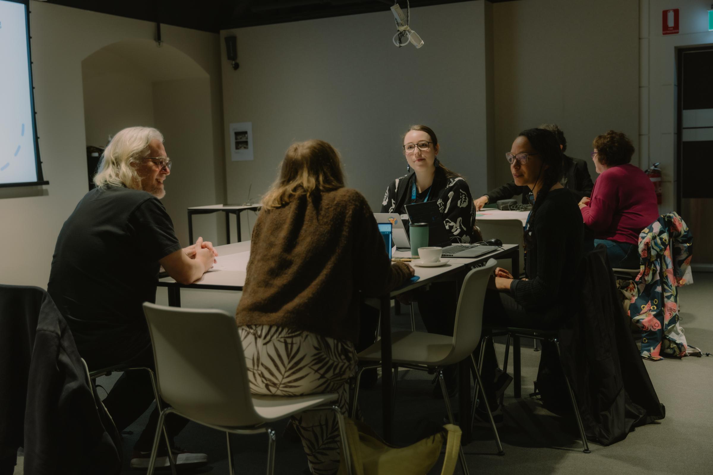 A man (Tim Sherratt) and 3 women seated at a table facing each other and conversing in a dimly lit room