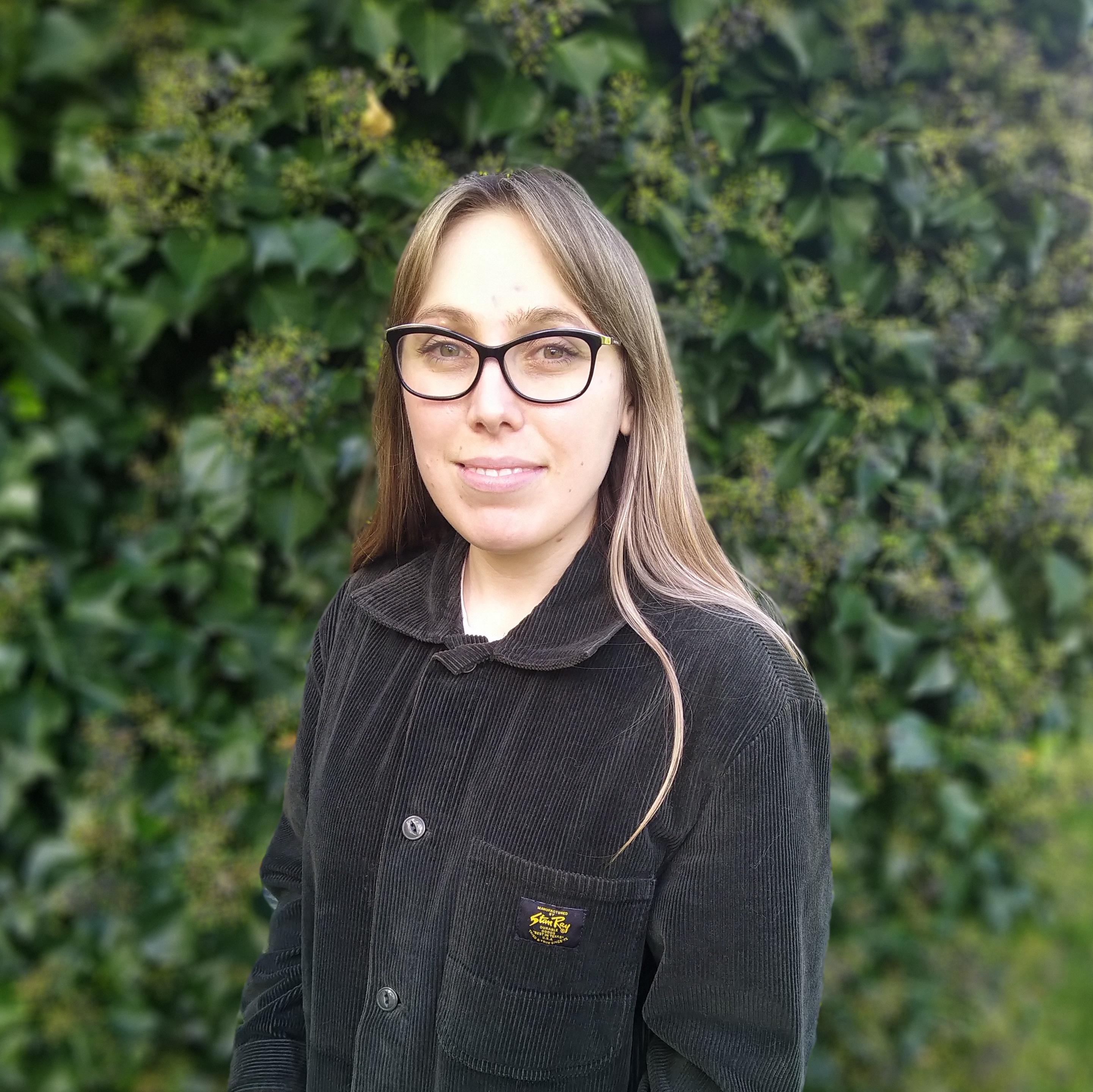 A headshot of a woman (Fiannuala Morgan) with long brown hair and thick black glasses against a leafy backdrop