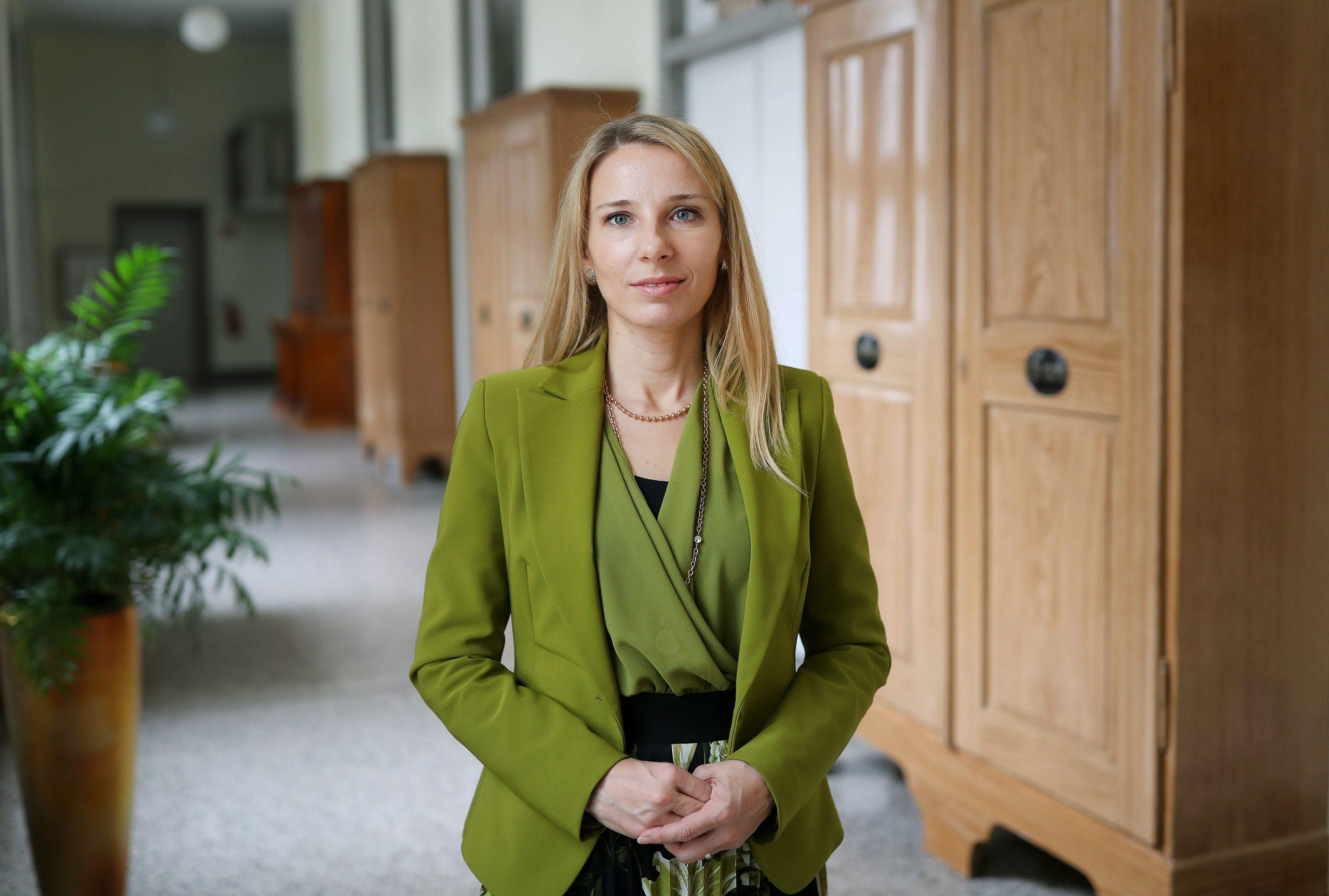 A woman wearing a green top with long blonde hair looking into the camera. She is in an academic setting