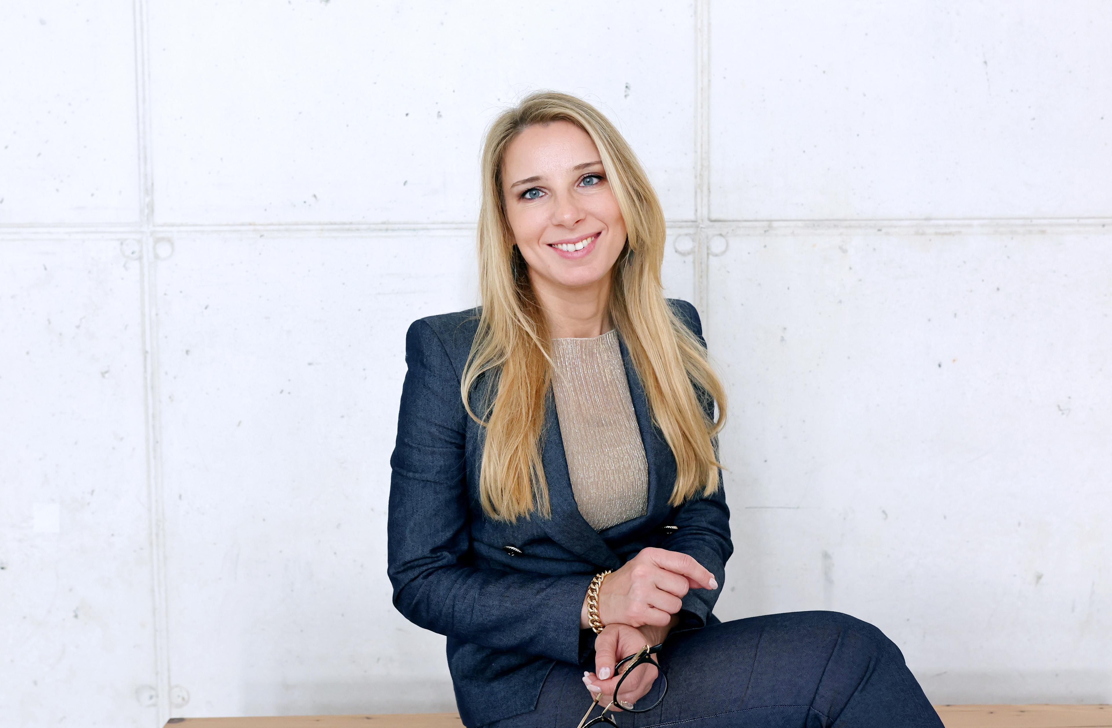 A woman (Ines Vodopivec) with long blonde hair, wearing a dark blue suit is seated and smiling at the camera