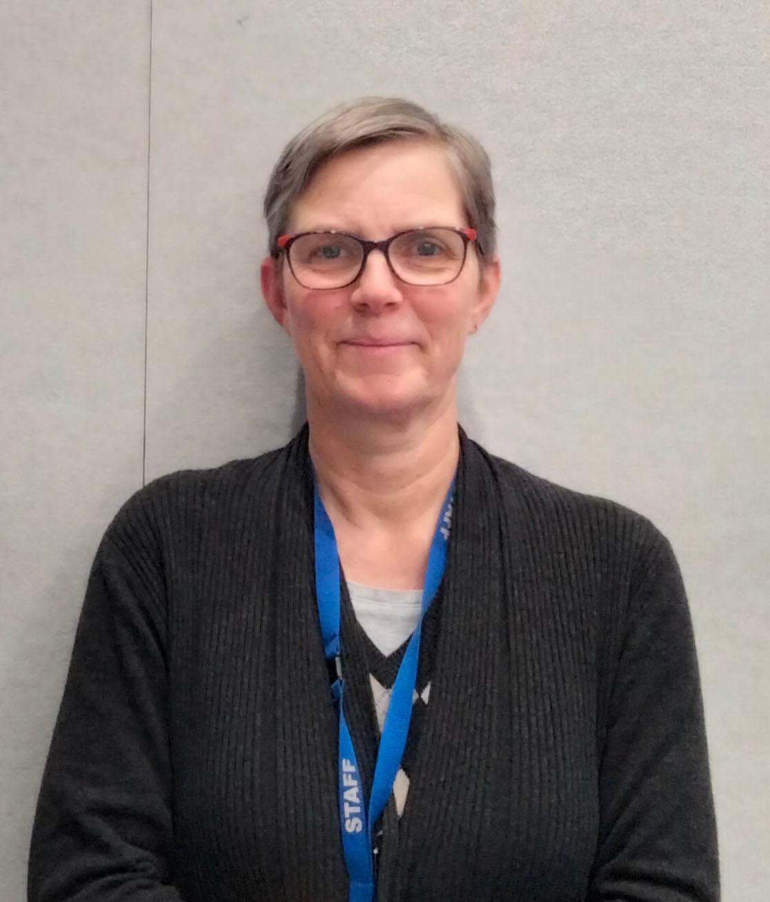 A portrait of woman (Suzie Geermans) with short brown hair and glasses looking at the camera with a slight smile wearing a blue lanyard around her neck