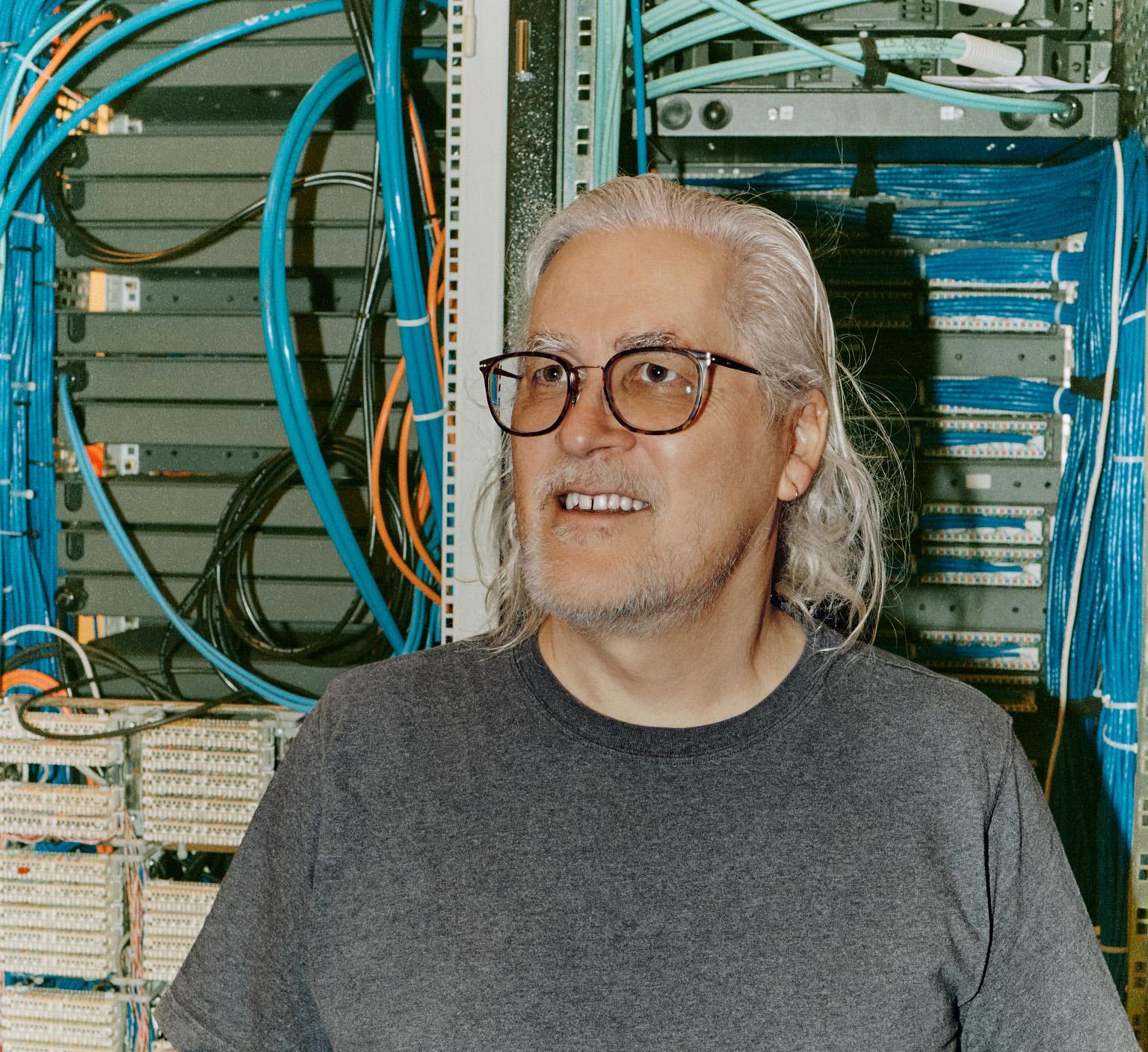 A cropped portrait of a man (Tim Sherratt) with long grey hair standing in front of computer server cables, wearing thick black glasses and a grey t-shirt