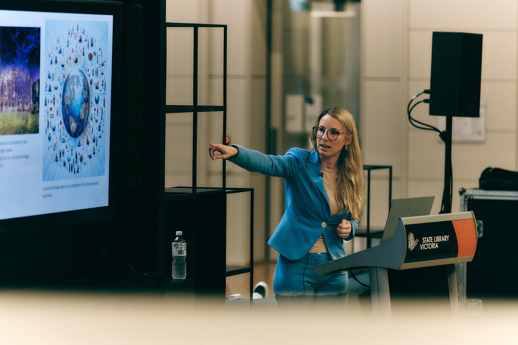 A woman (Ines Vodopivec), standing in front of a lectern, wearing glasses and a bright blue suit points at a presentation screen