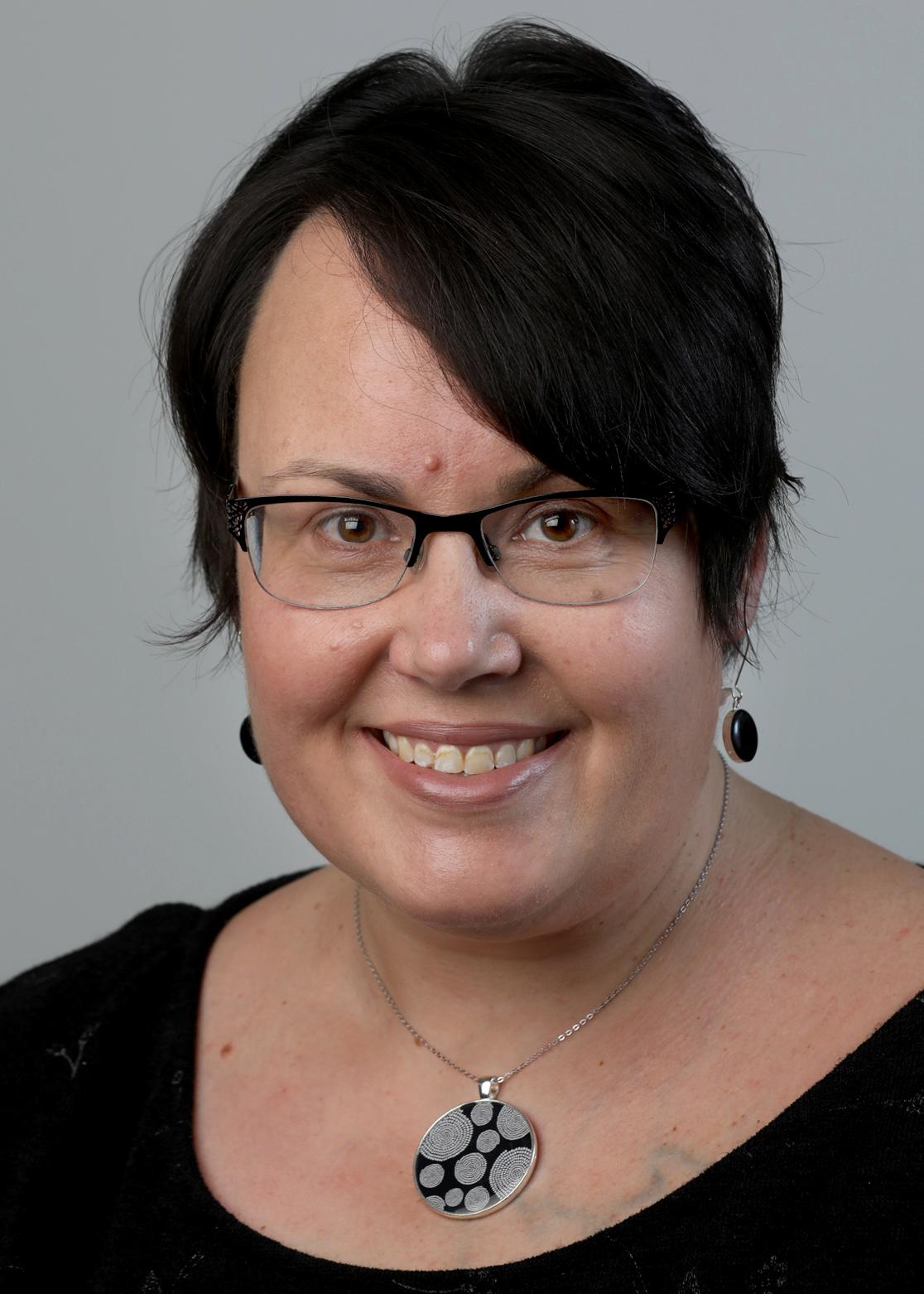 A portrait of a smiling woman with short black hair and glasses. She has black earrings and a silver necklace